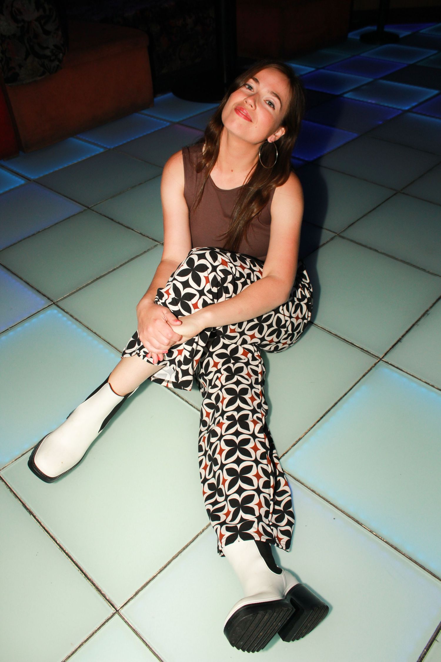Young woman sitting on illuminated dance floor wearing floral pants, brown tank top, and white boots.