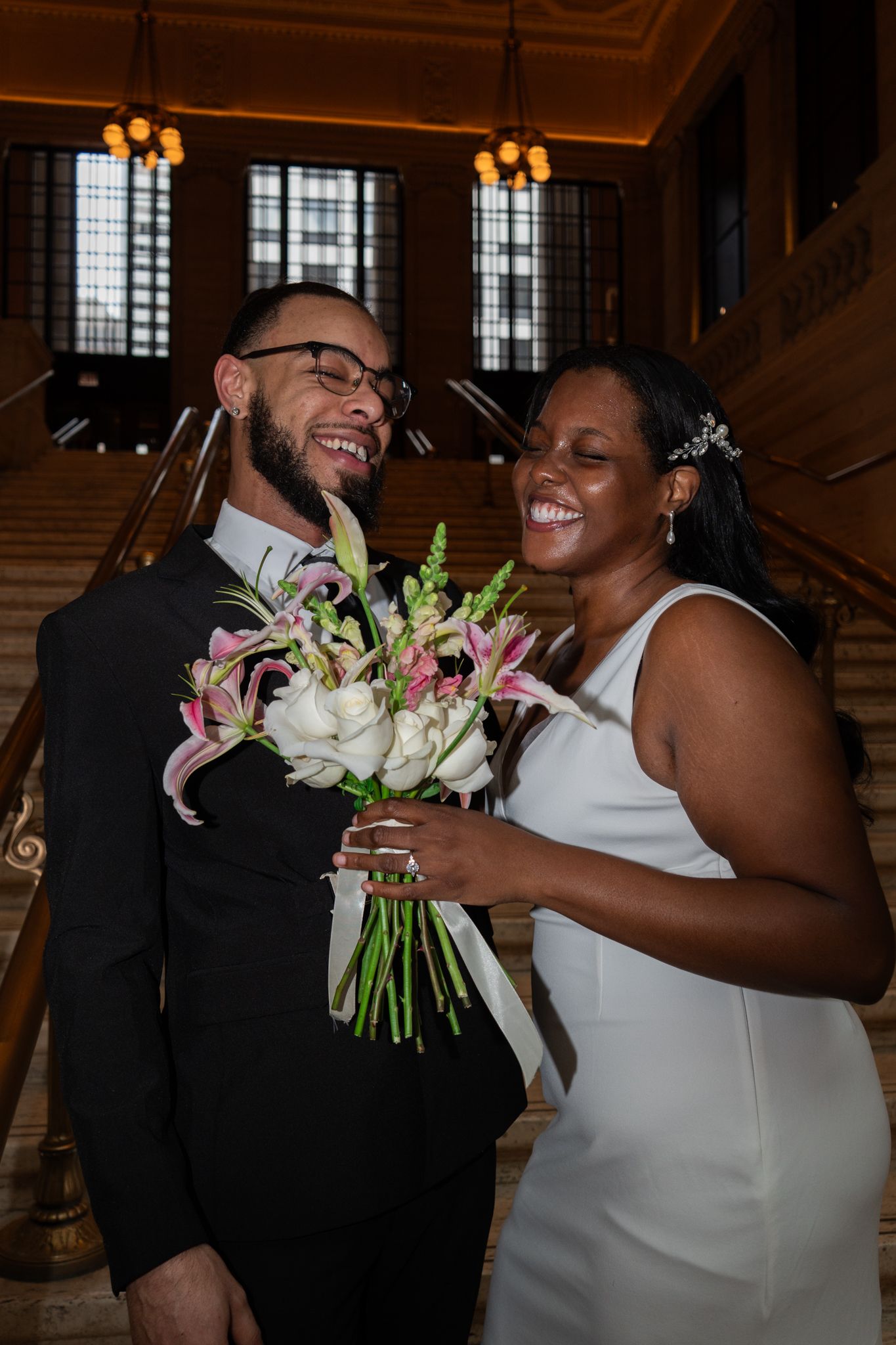 Joyful newlywed couple holding pink and white bouquet laughing on grand staircase at elegant venue.