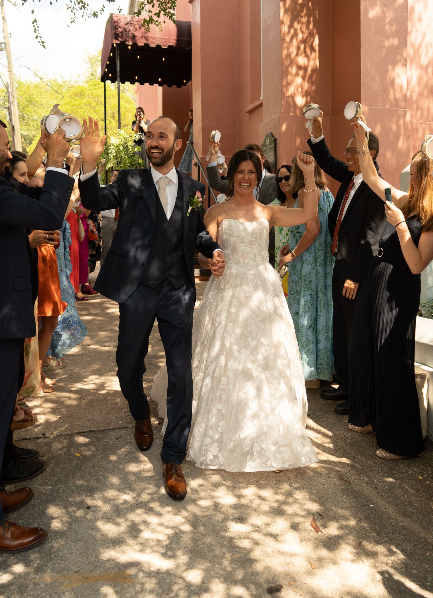 Joyful newlyweds walk through a celebratory sendoff with guests raising hands outside a pink building.