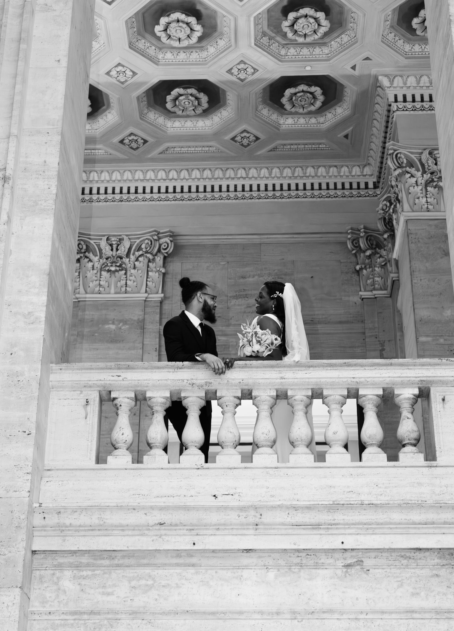 Black and white photo of a bride and groom standing on an ornate marble balcony with decorative ceiling.