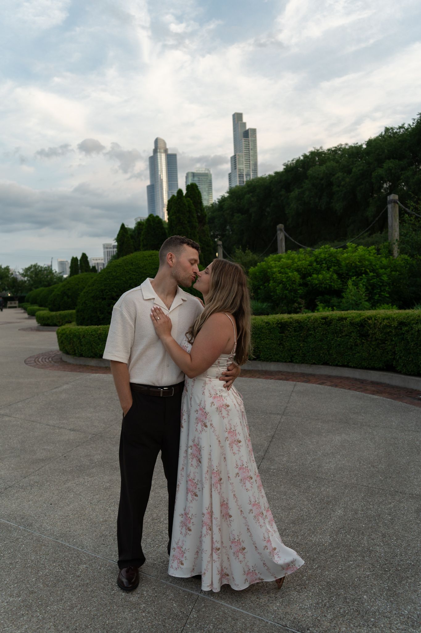 Couple sharing a romantic kiss in an urban park with lush greenery and city skyscrapers in the background.