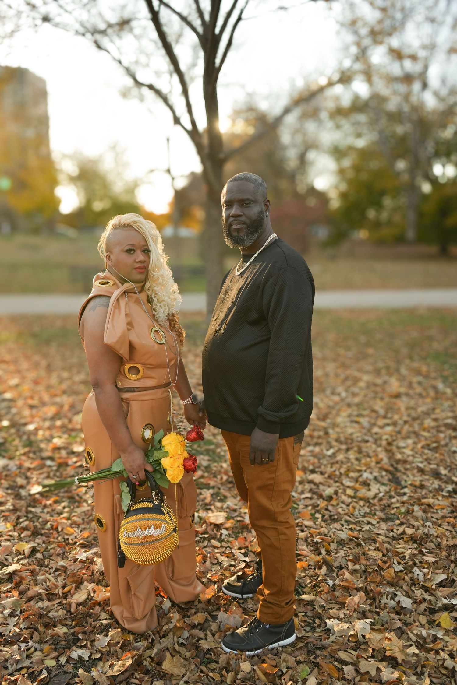Couple posing in autumn park, woman in tan outfit holding flowers, man in dark green sweater and gold pants.