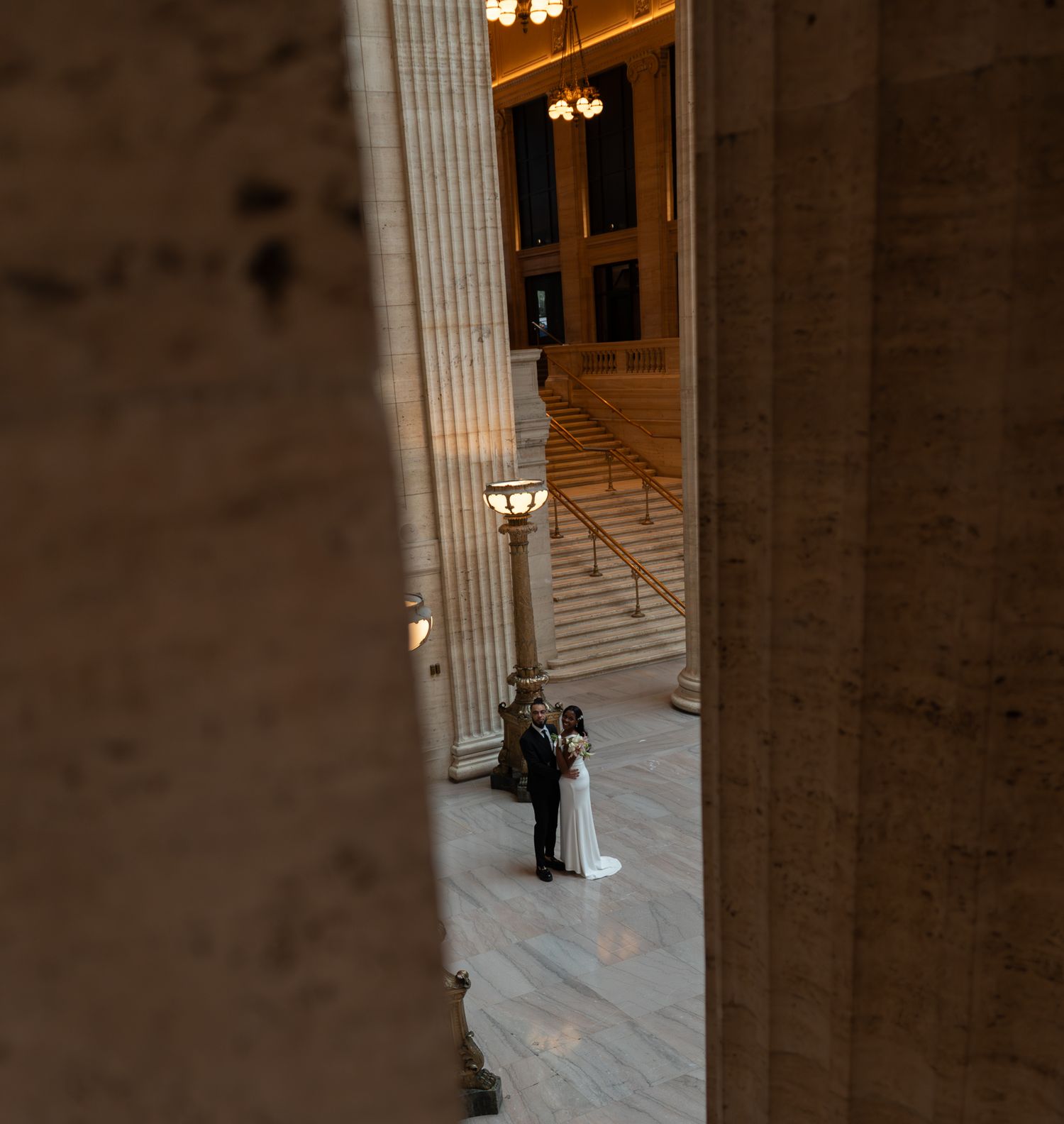 Bride and groom share a romantic moment inside a grand marble building with tall columns and chandeliers.