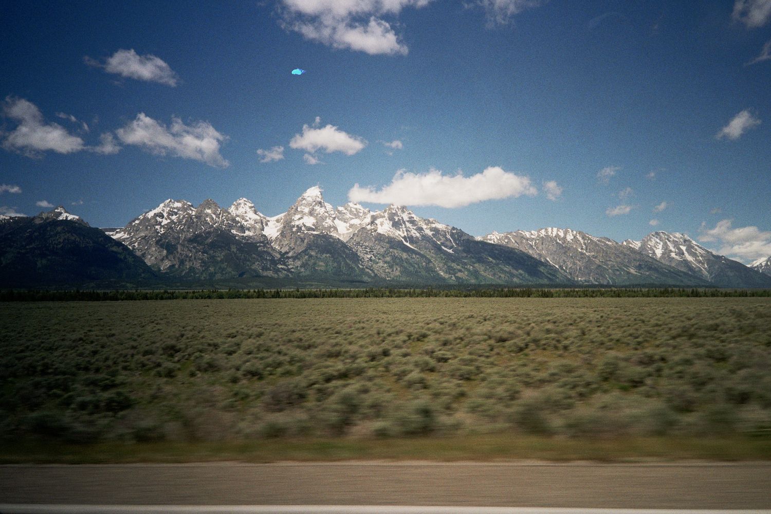 Snow-capped Grand Teton mountain range under blue sky with green plains in foreground, Wyoming landscape.