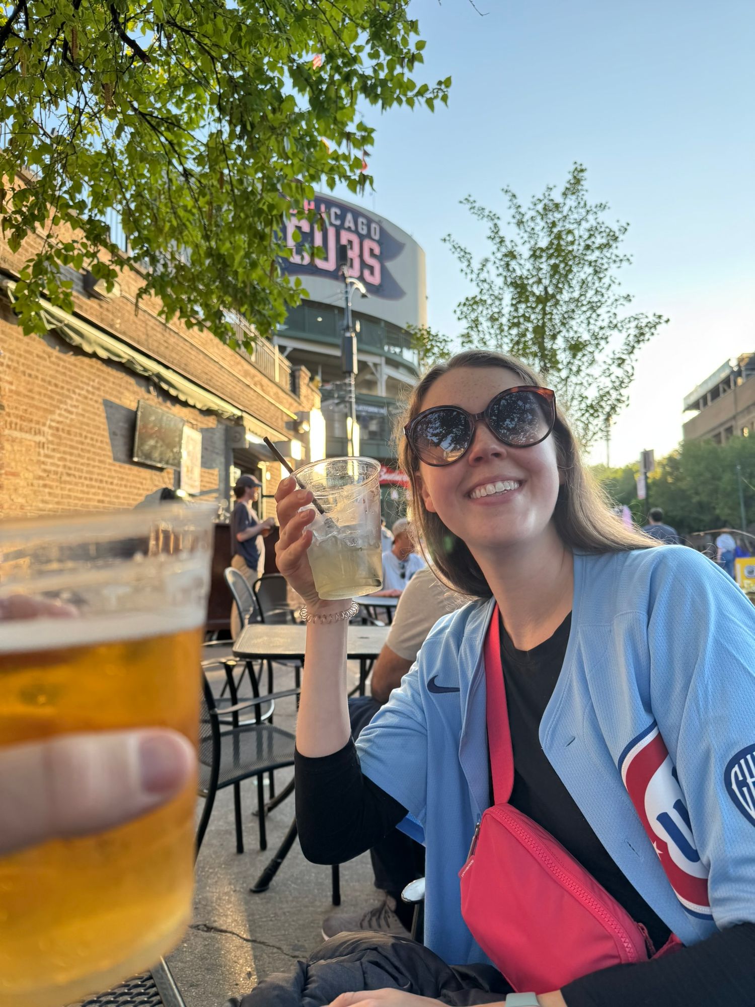 Woman in Chicago Cubs jersey toasting drinks at outdoor bar patio near Wrigley Field on sunny day | Sydney Allen Photography