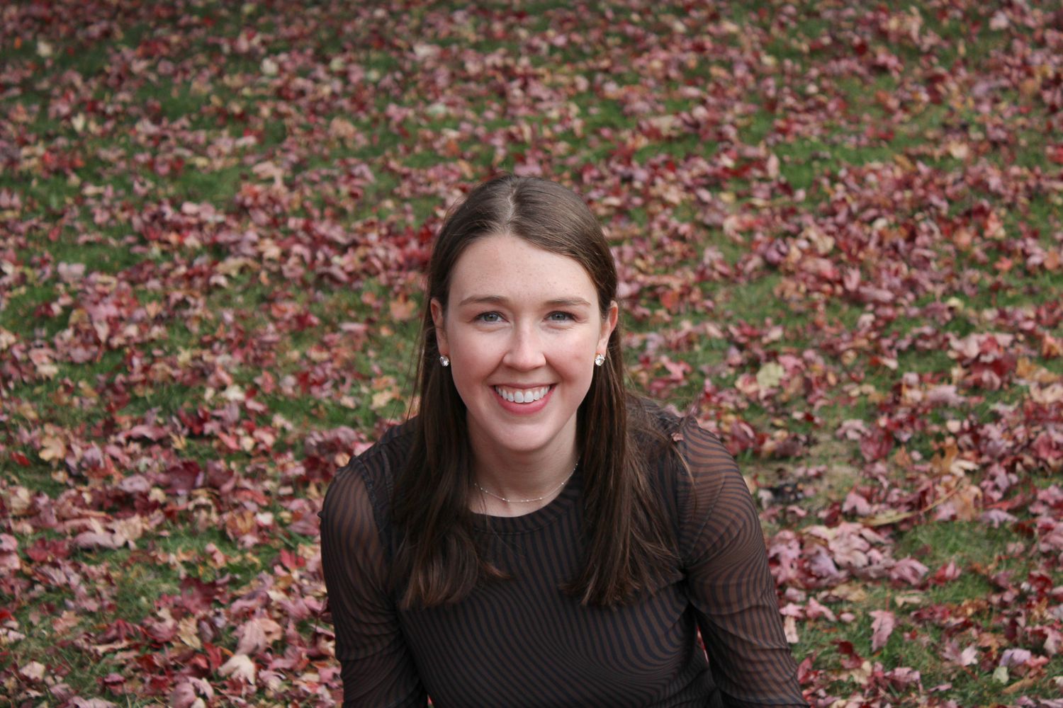 Smiling young woman sitting outdoors among fallen autumn red leaves on green grass.