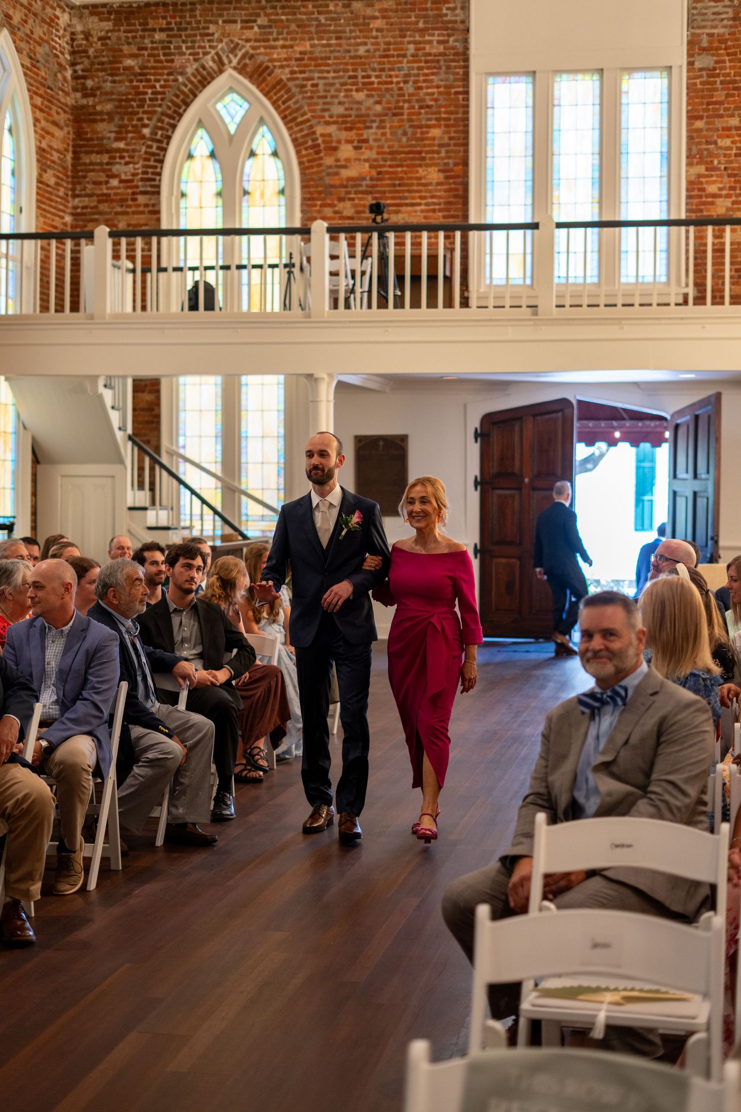 Elegant wedding processional in a historic brick church with guests seated in white chairs lining the aisle.