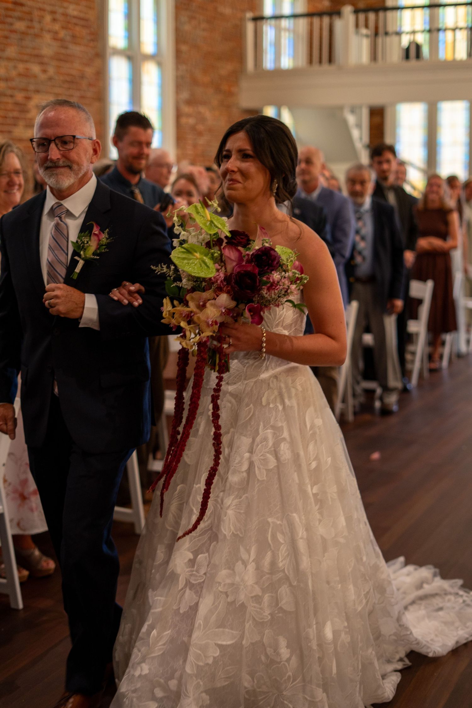 Emotional bride in lace gown walks down aisle with escort, holding burgundy bouquet in elegant brick wedding venue.