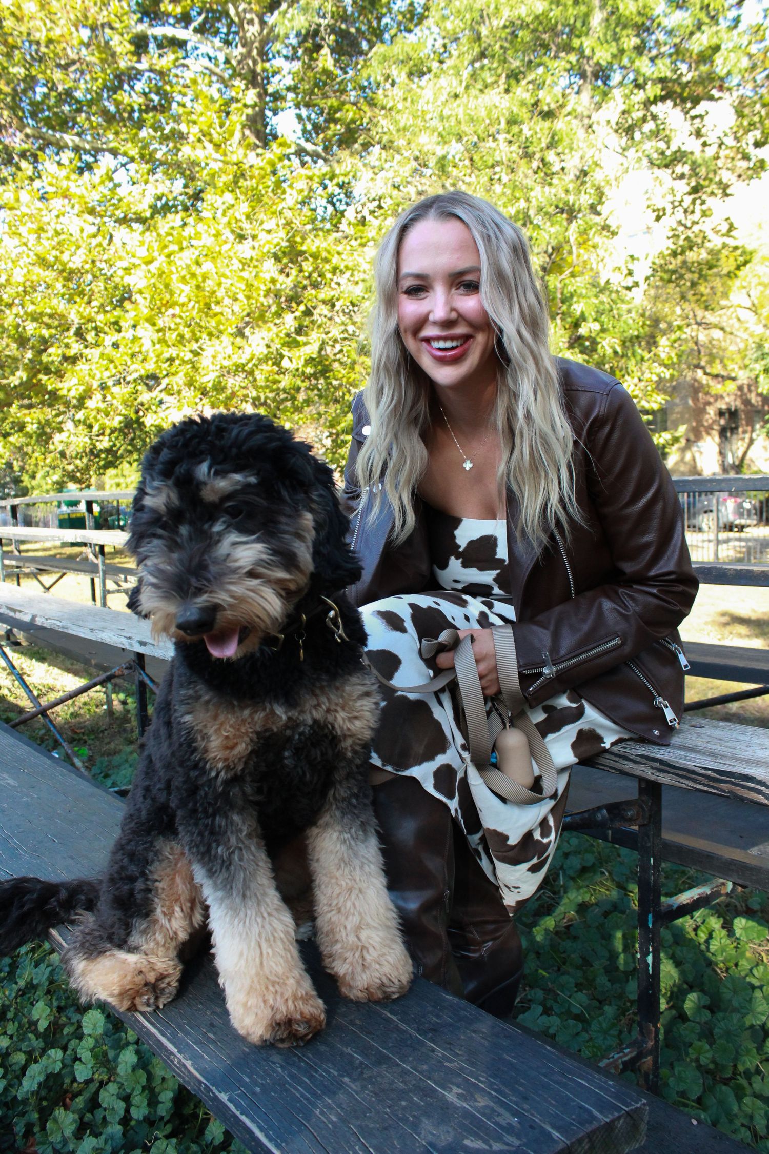 Smiling blonde woman in brown leather jacket and cow print pants sitting on wooden bench with large Airedale Terrier dog outdoors.
