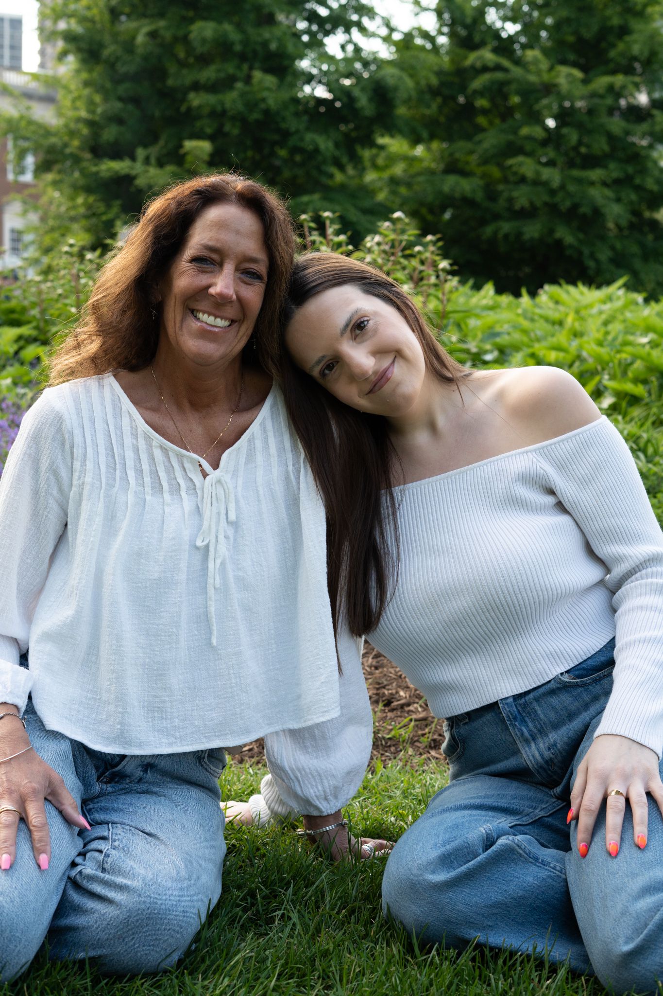 Two women in white tops and jeans sit together on grass, smiling, with lush green garden in background.