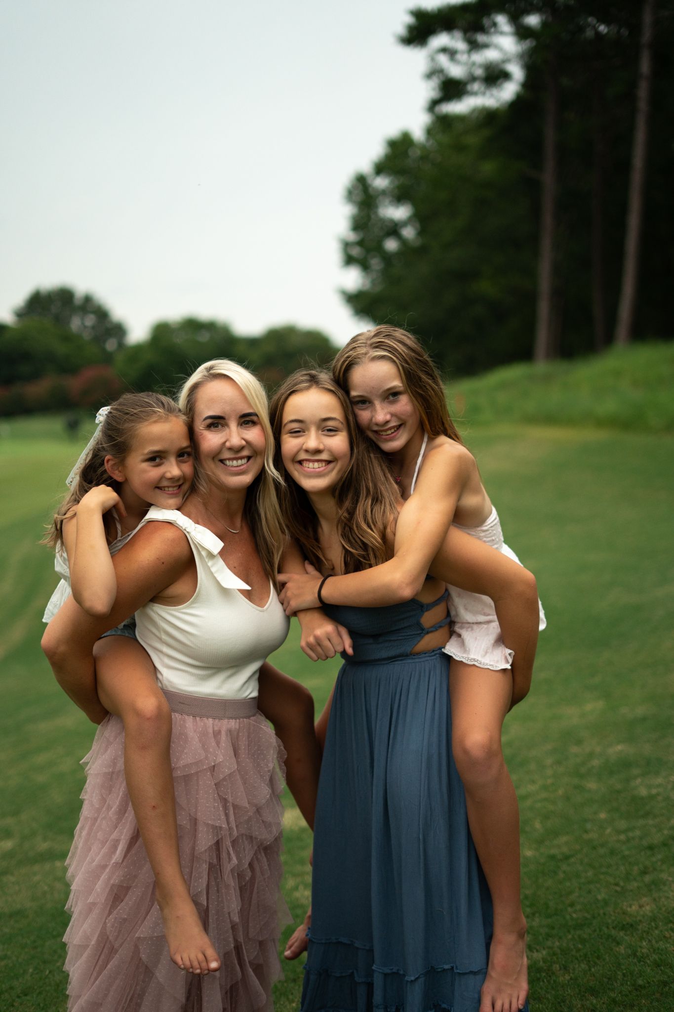 Four smiling females pose together outdoors on lush green grass with tall trees in background.