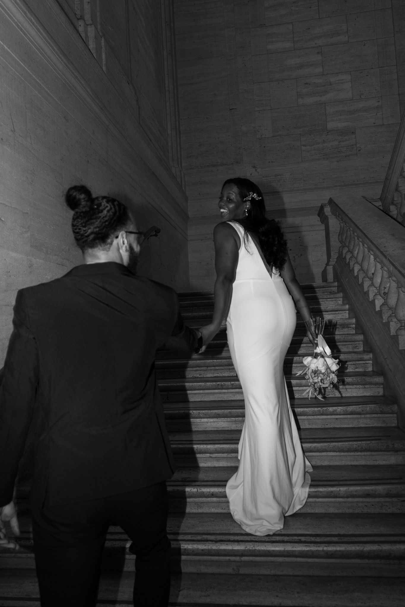 Black and white photo of a bride in white gown and groom in suit walking up grand stone staircase.