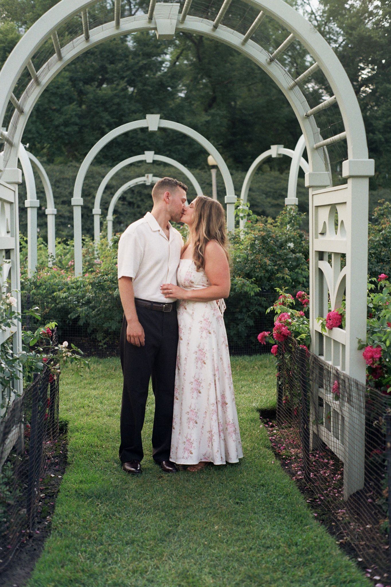 Couple sharing a romantic kiss under white garden arbor arches surrounded by lush greenery and blooming roses.