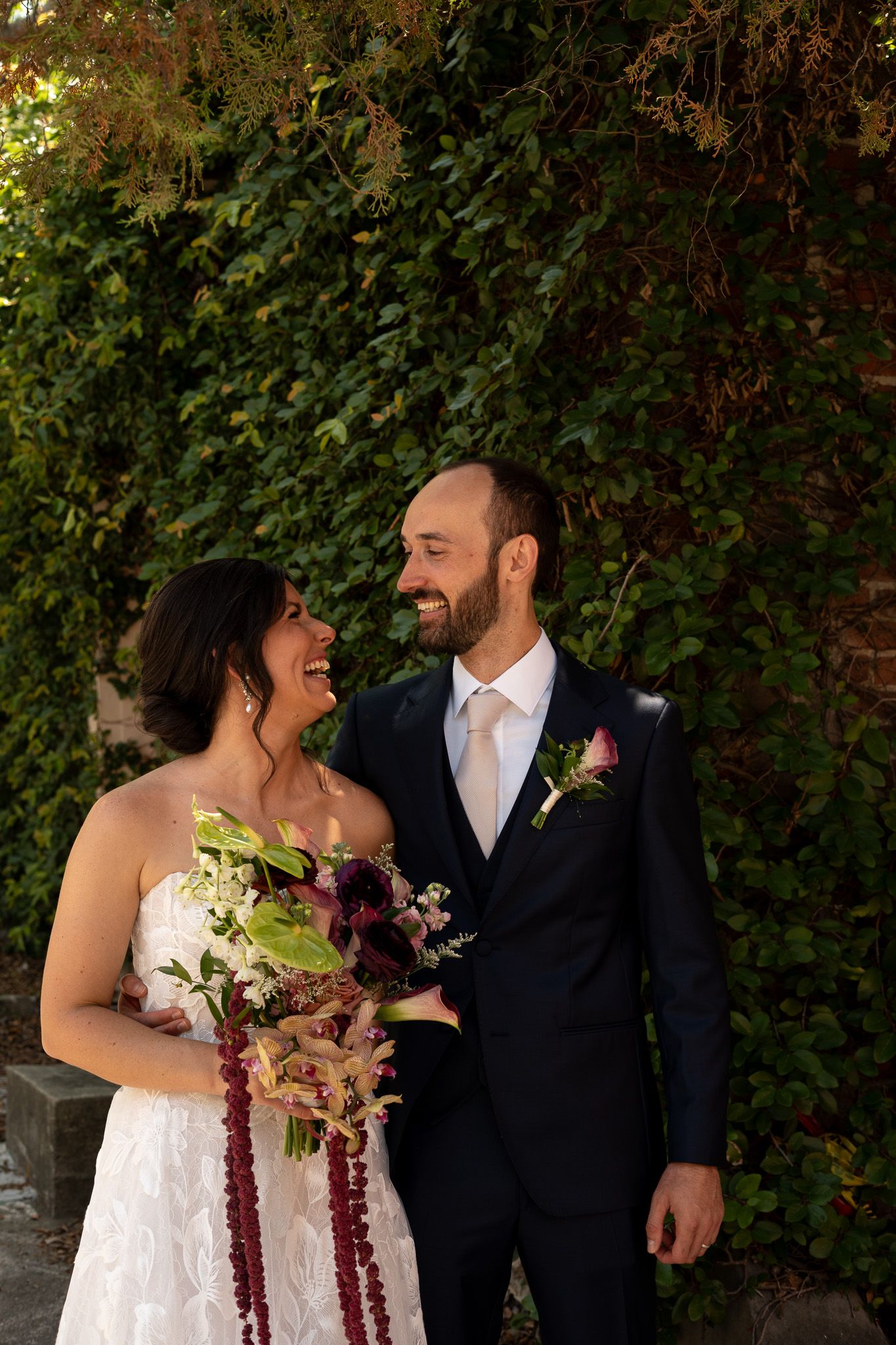 A bride and groom share a joyful moment outdoors, she holds a burgundy bouquet, he wears a navy suit with floral boutonniere.