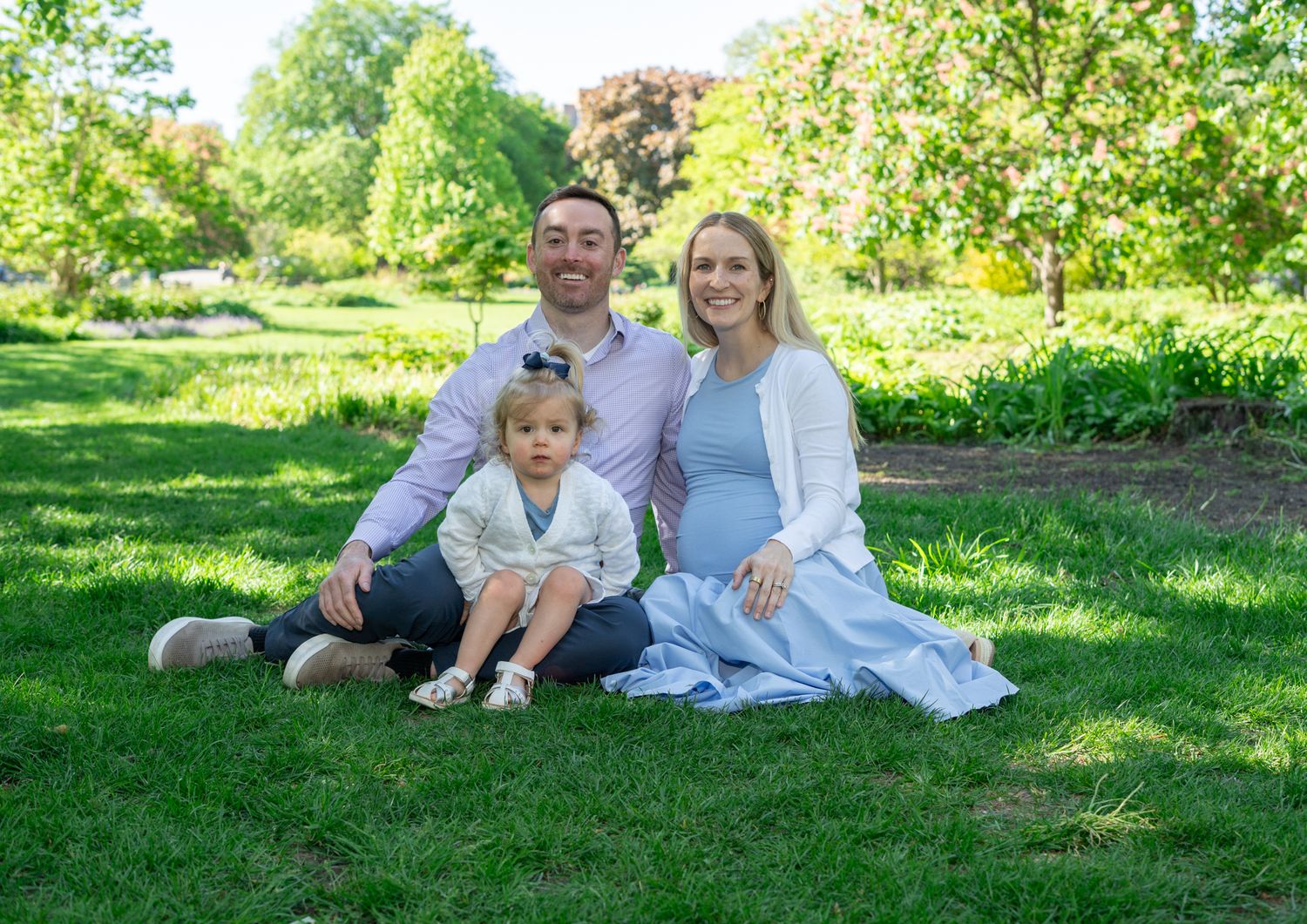 Happy family of three sitting on green grass in a sunny park, pregnant mother in white cardigan.