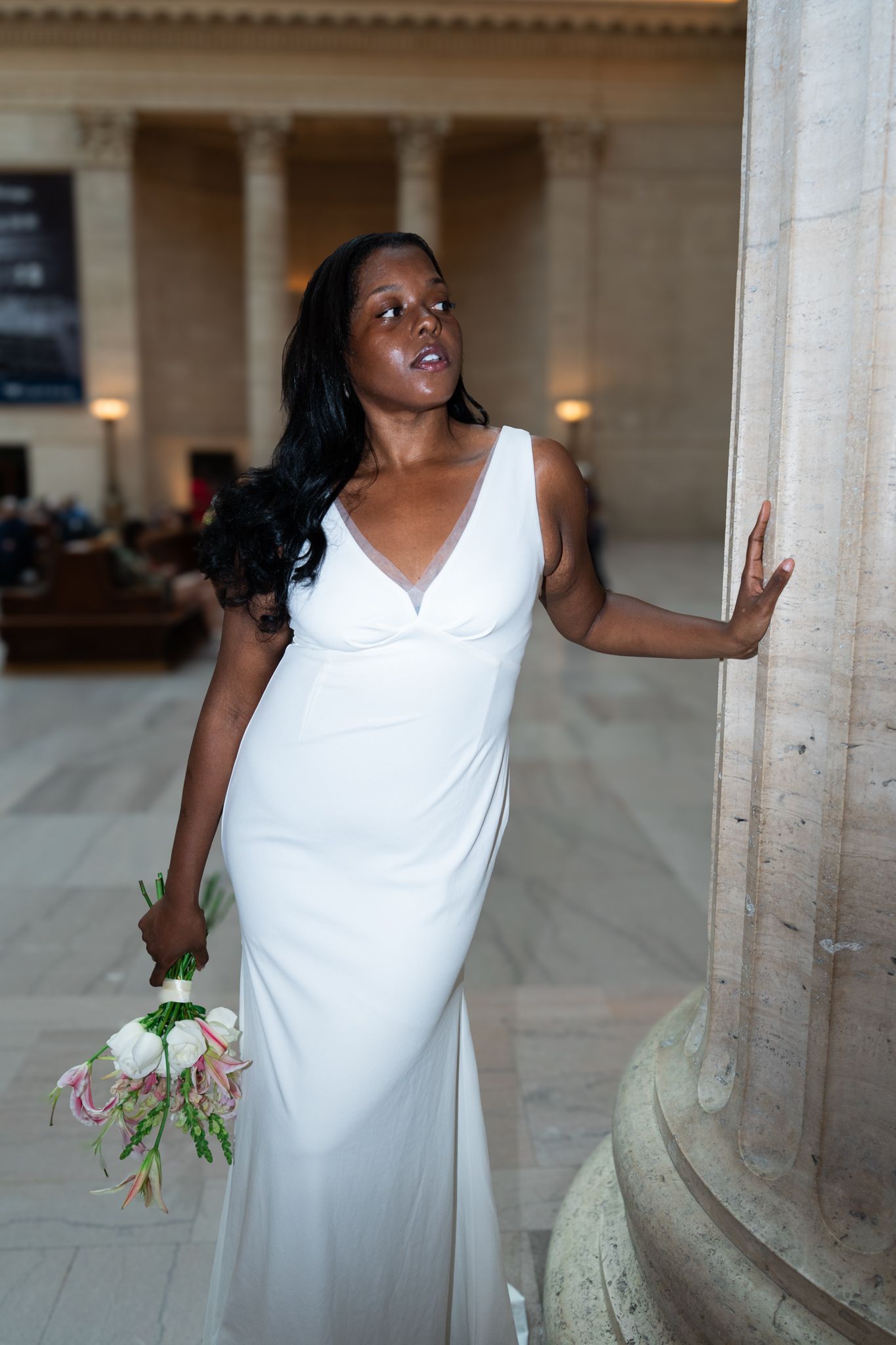 Bride in white gown holding bouquet leans against marble column in elegant hall.