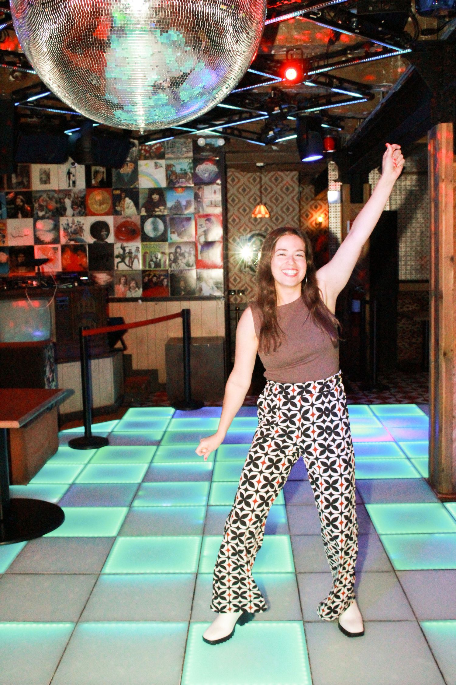 Woman dancing under a disco ball on a lit dance floor in a retro-themed venue.