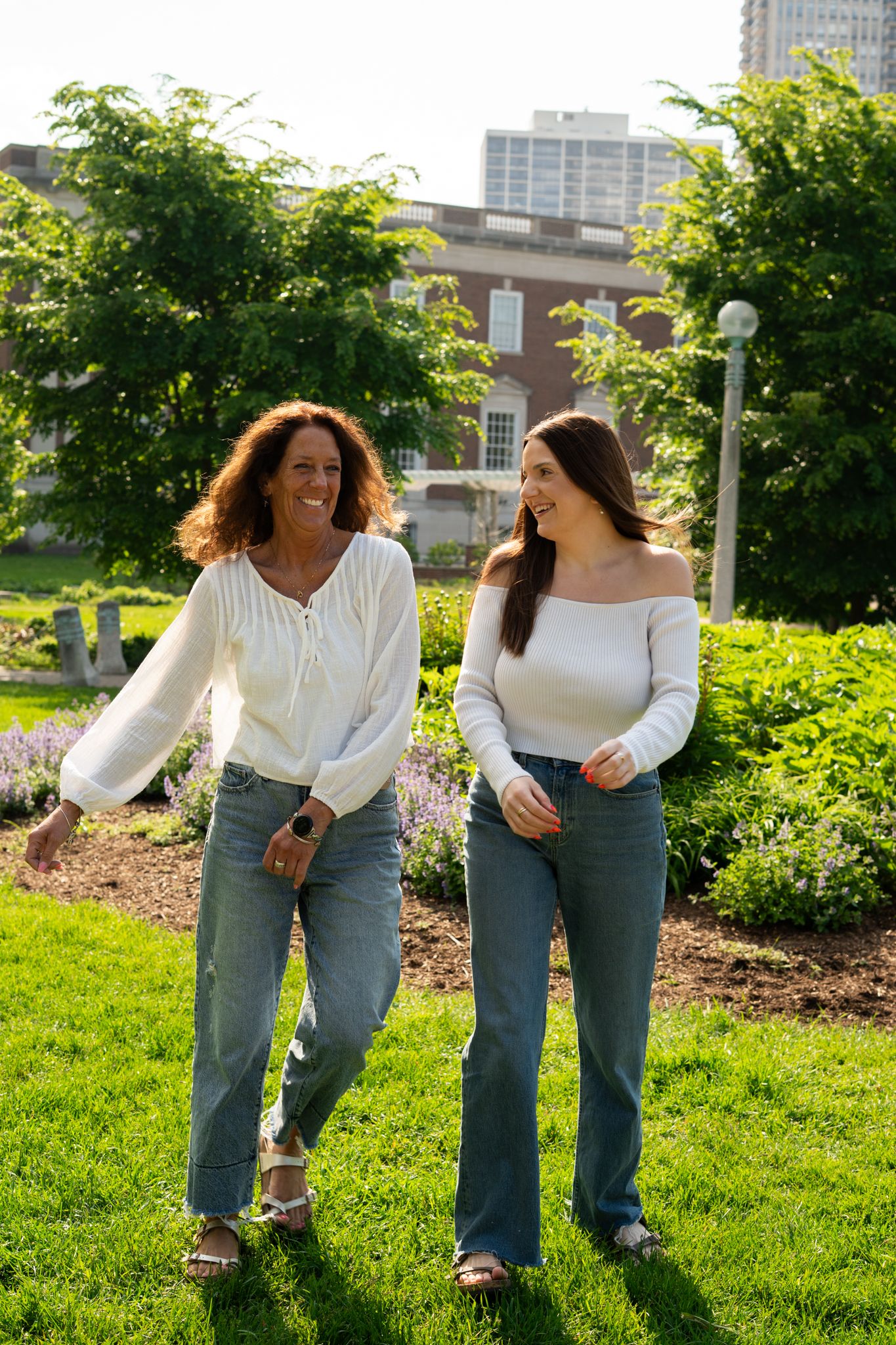 Two women in white tops and jeans walking together in a lush green park on a sunny day.