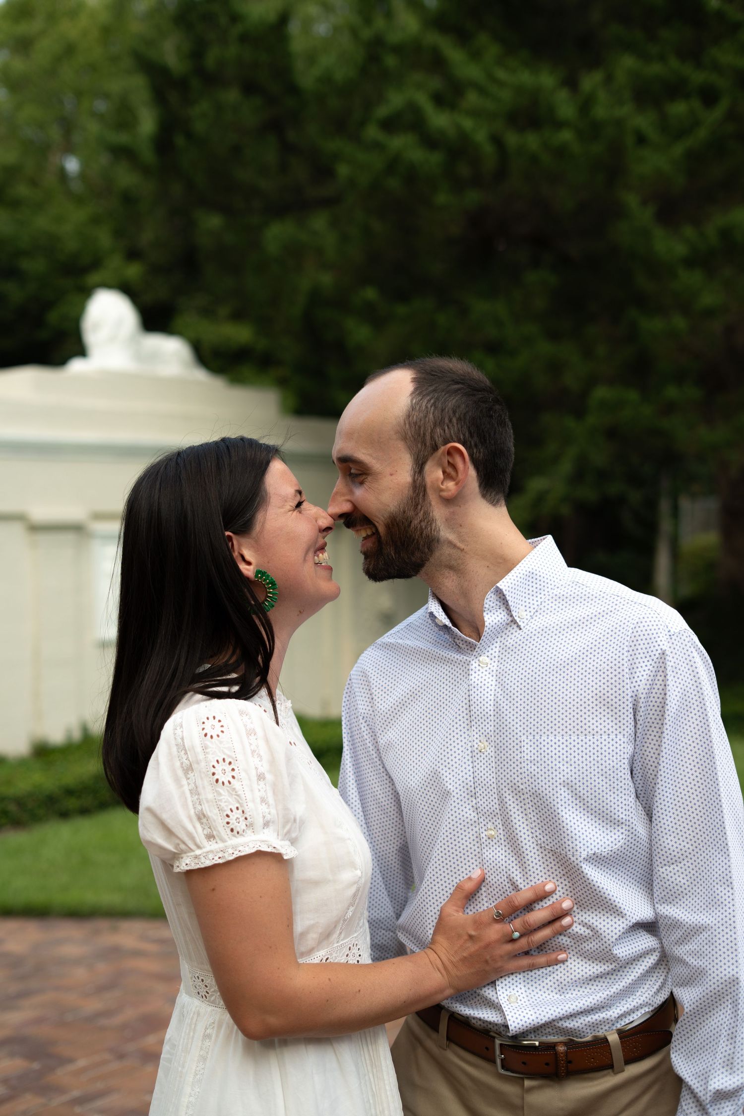 A couple shares a romantic nose-to-nose moment outdoors, woman in white dress with green earrings, man in light blue shirt.
