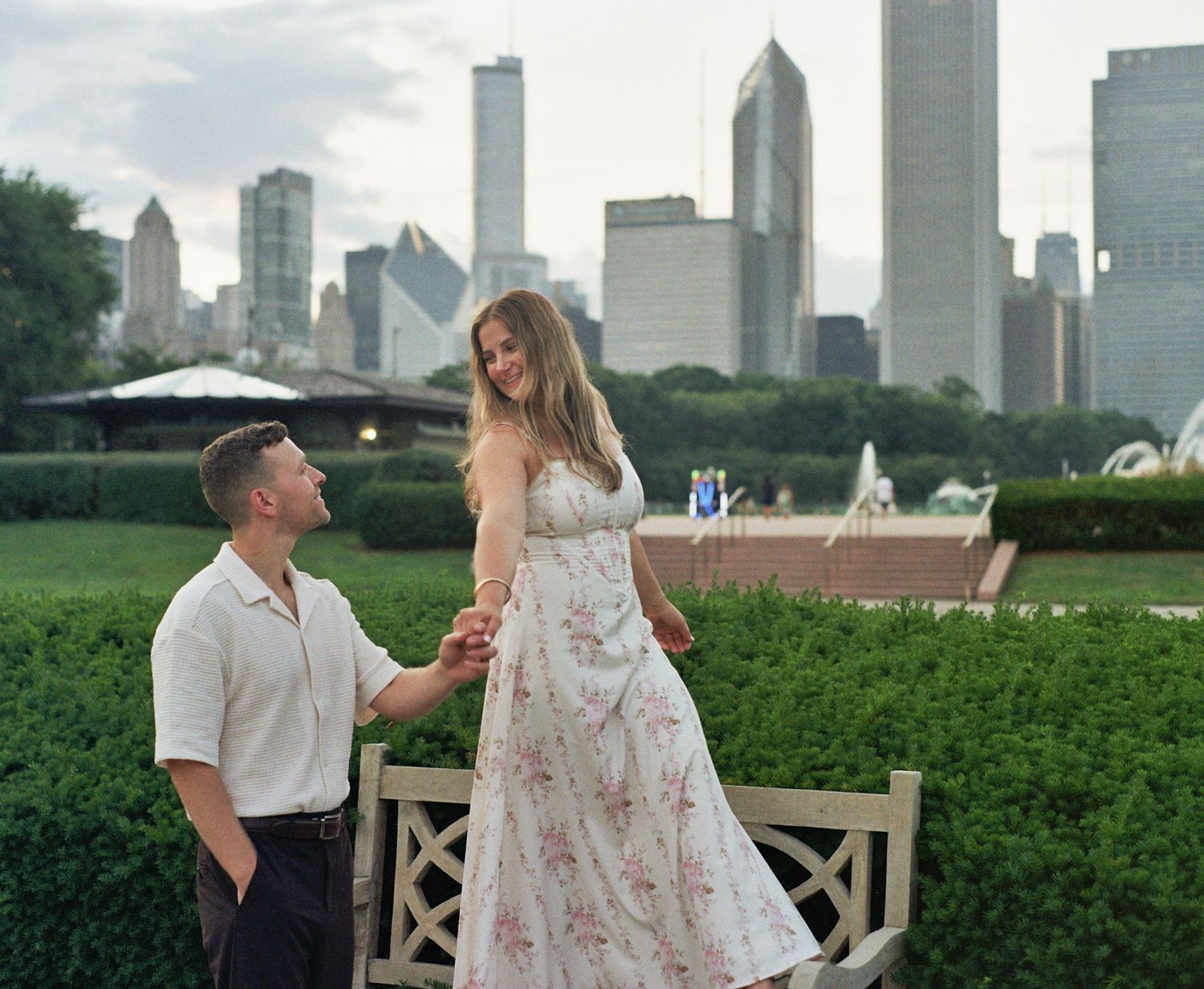 Couple holding hands in a garden with Chicago skyline in the background, woman in floral dress standing on bench.