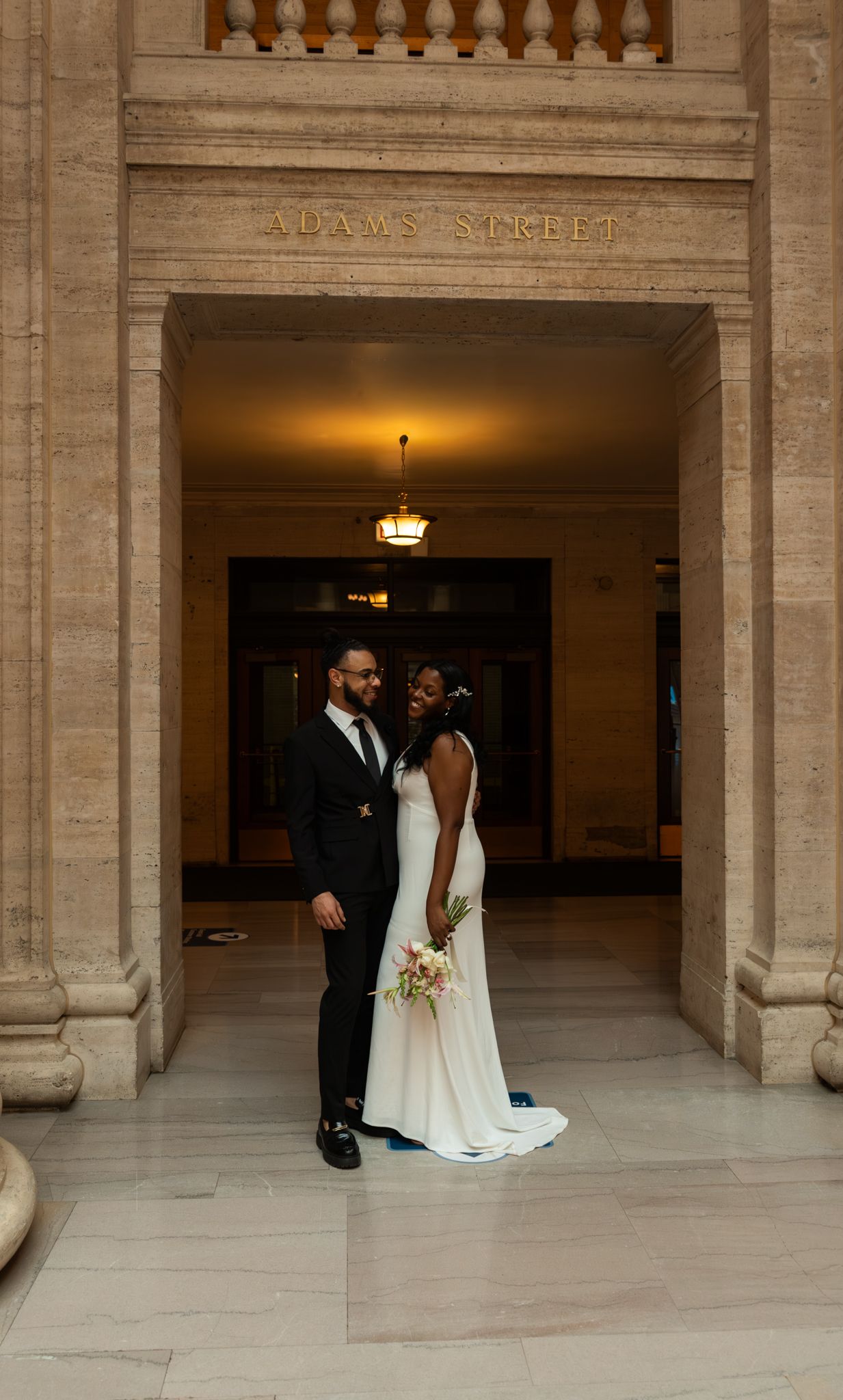 Bride and groom pose under Adams Street archway in elegant wedding attire at a grand stone building.