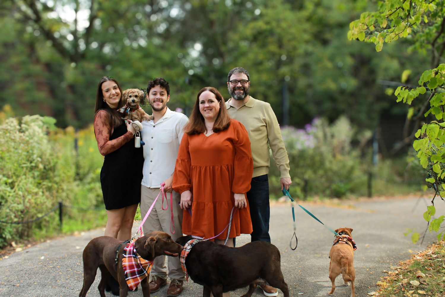 Family of four smiling outdoors on a path with three dogs, surrounded by lush green trees.
