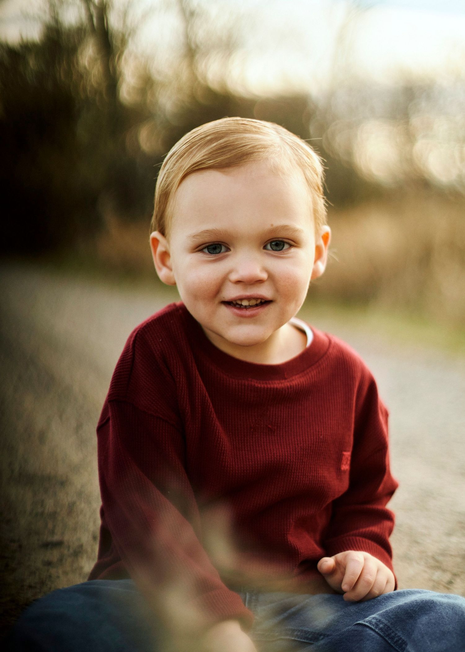 Child in burgundy sweater smiling during outdoor autumn photoshoot.