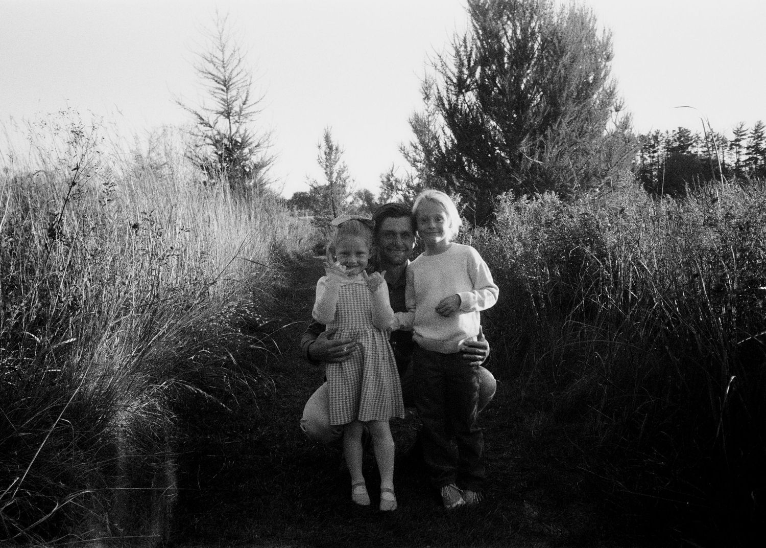 Black and white photo of family standing among tall grass and pine trees.