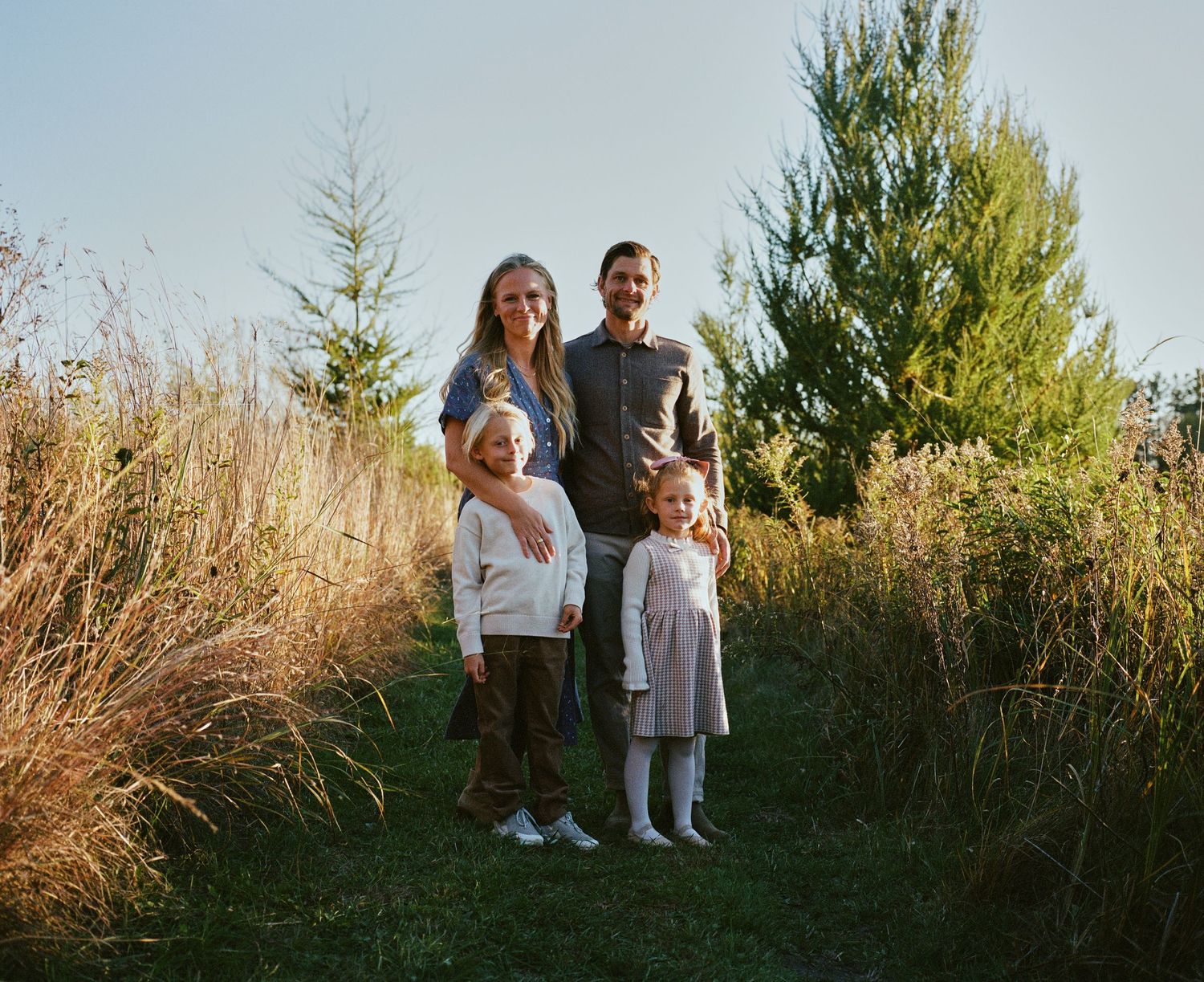 Family standing together among evergreen trees during outdoor photo session at sunset.