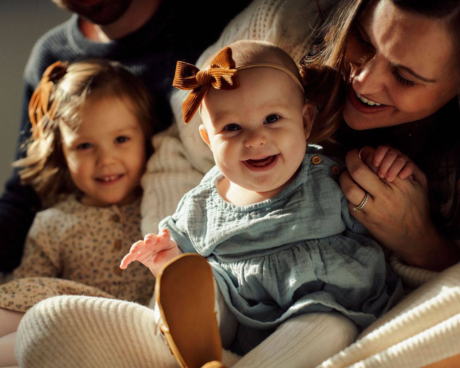 A baby wearing a blue dress and orange bow sits with family members on a cozy white blanket in warm lighting.