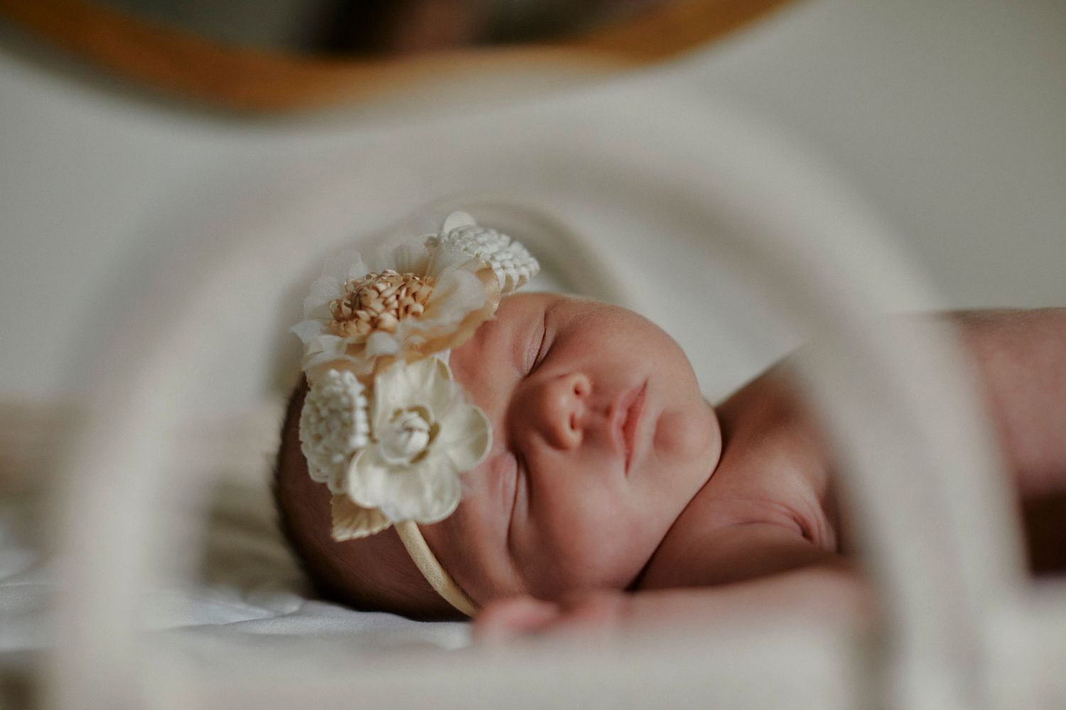 Peaceful newborn baby wearing an ornate floral headband resting on white bedding.