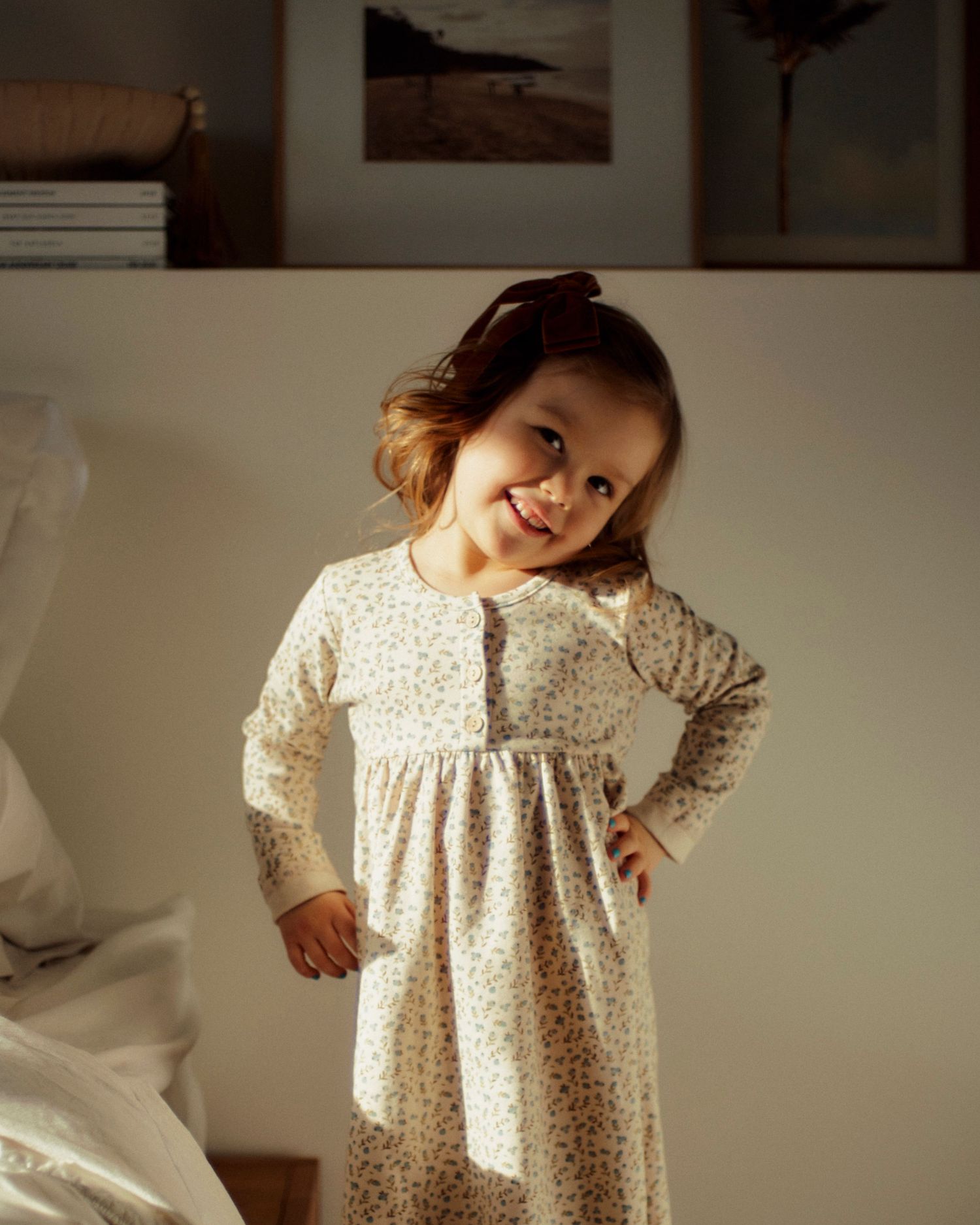 A young child wearing a floral print empire waist dress stands in a warmly lit room with framed artwork on the wall.