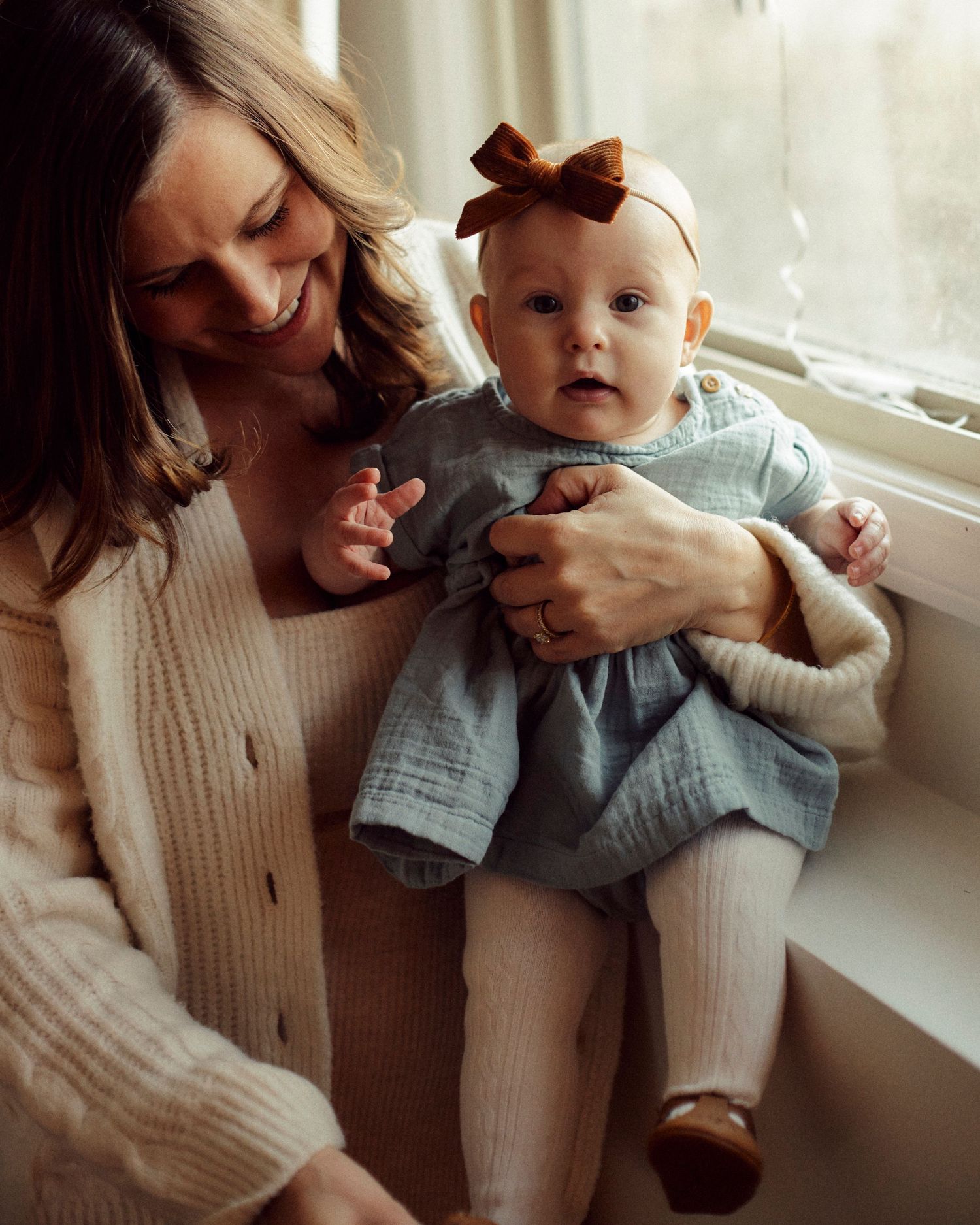 A tender moment by the window shows a baby in a blue dress with a bow sitting on a caregiver's lap in a cozy sweater.
