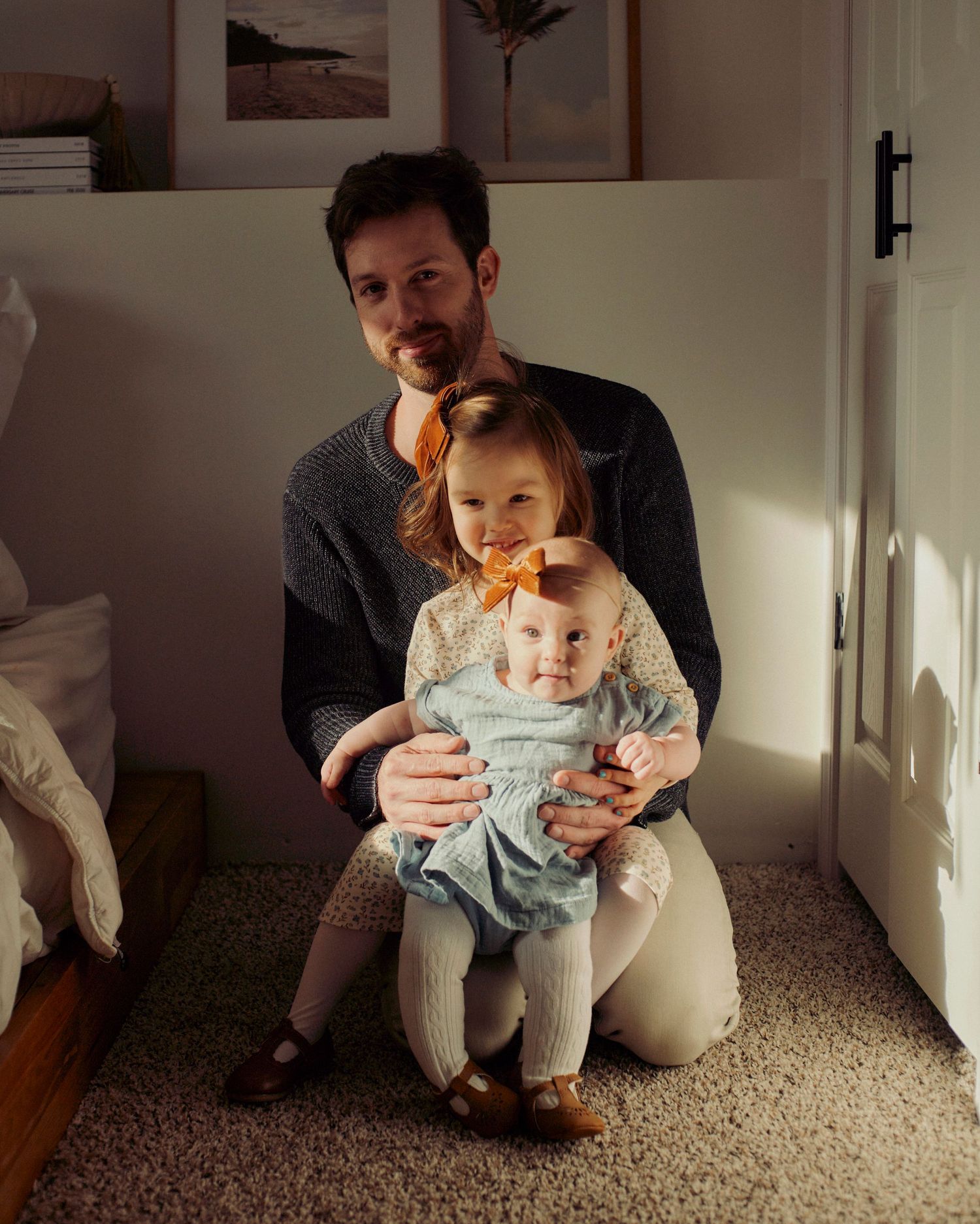 A parent sits on the floor holding two small children in a warmly lit room with framed artwork on the wall.