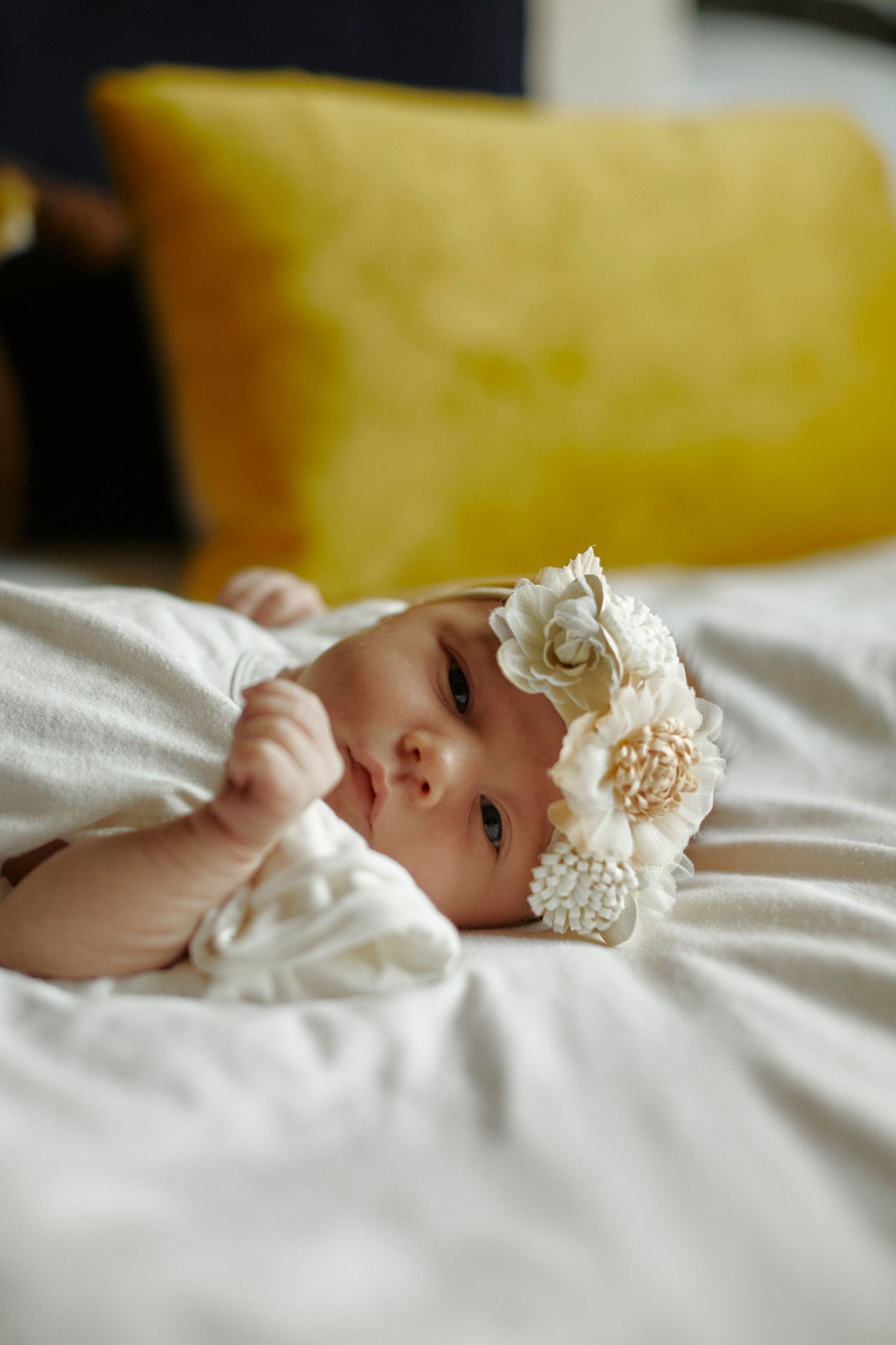 Baby wearing a white dress and floral headband lying on white bedding next to a yellow pillow.