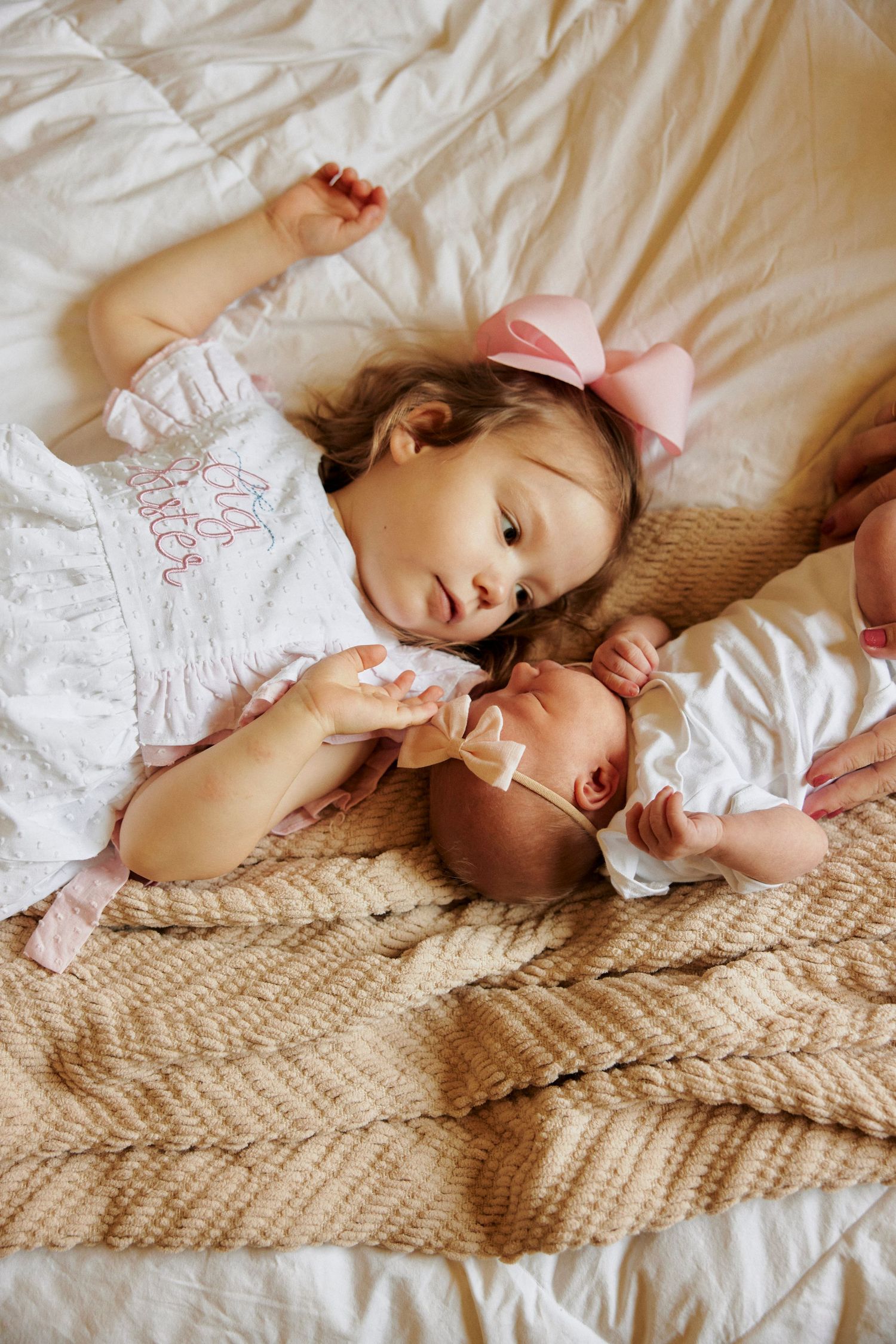Two children lying together on a textured blanket wearing white and pink outfits.