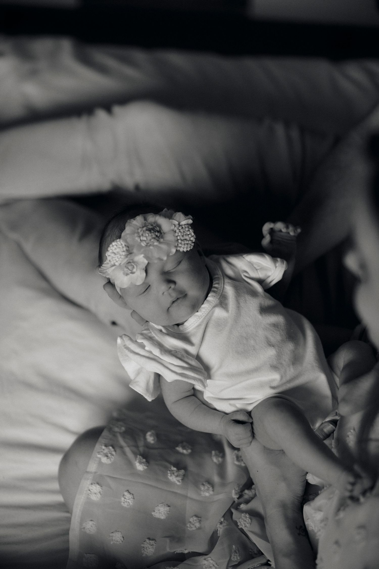 Black and white portrait of a sleeping infant wearing a lace headband on soft bedding.