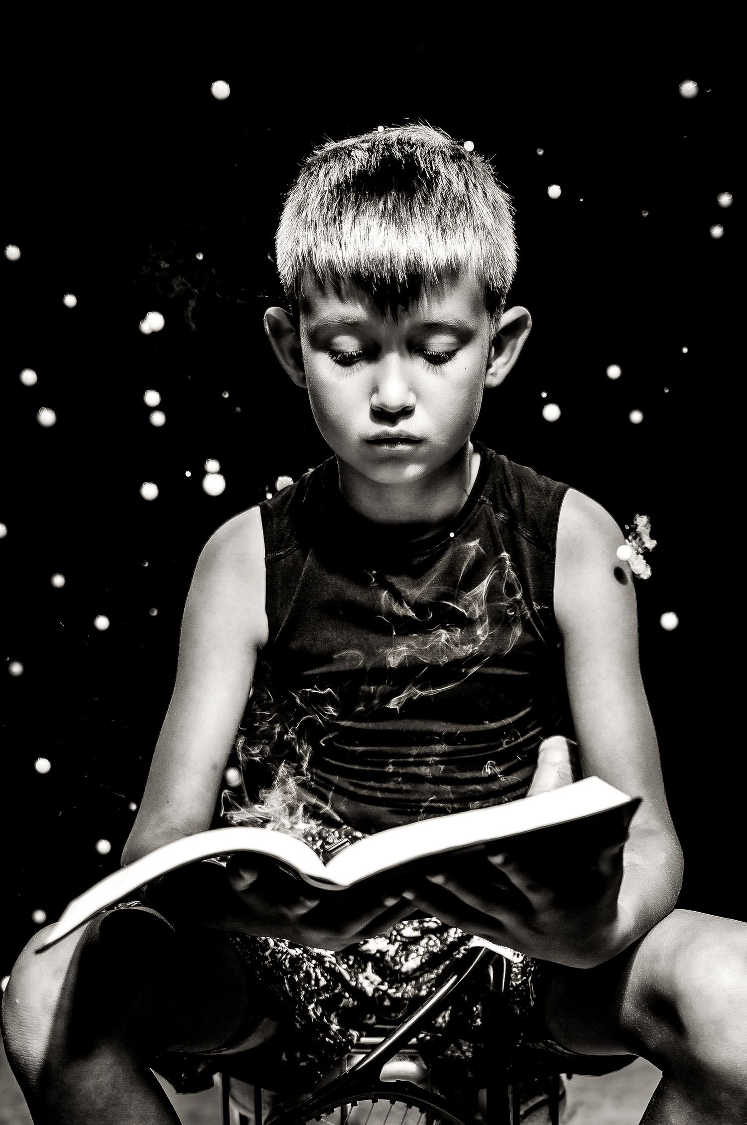 A young boy sitting on a chair and reading a book in a dark setting with sparkling lights.