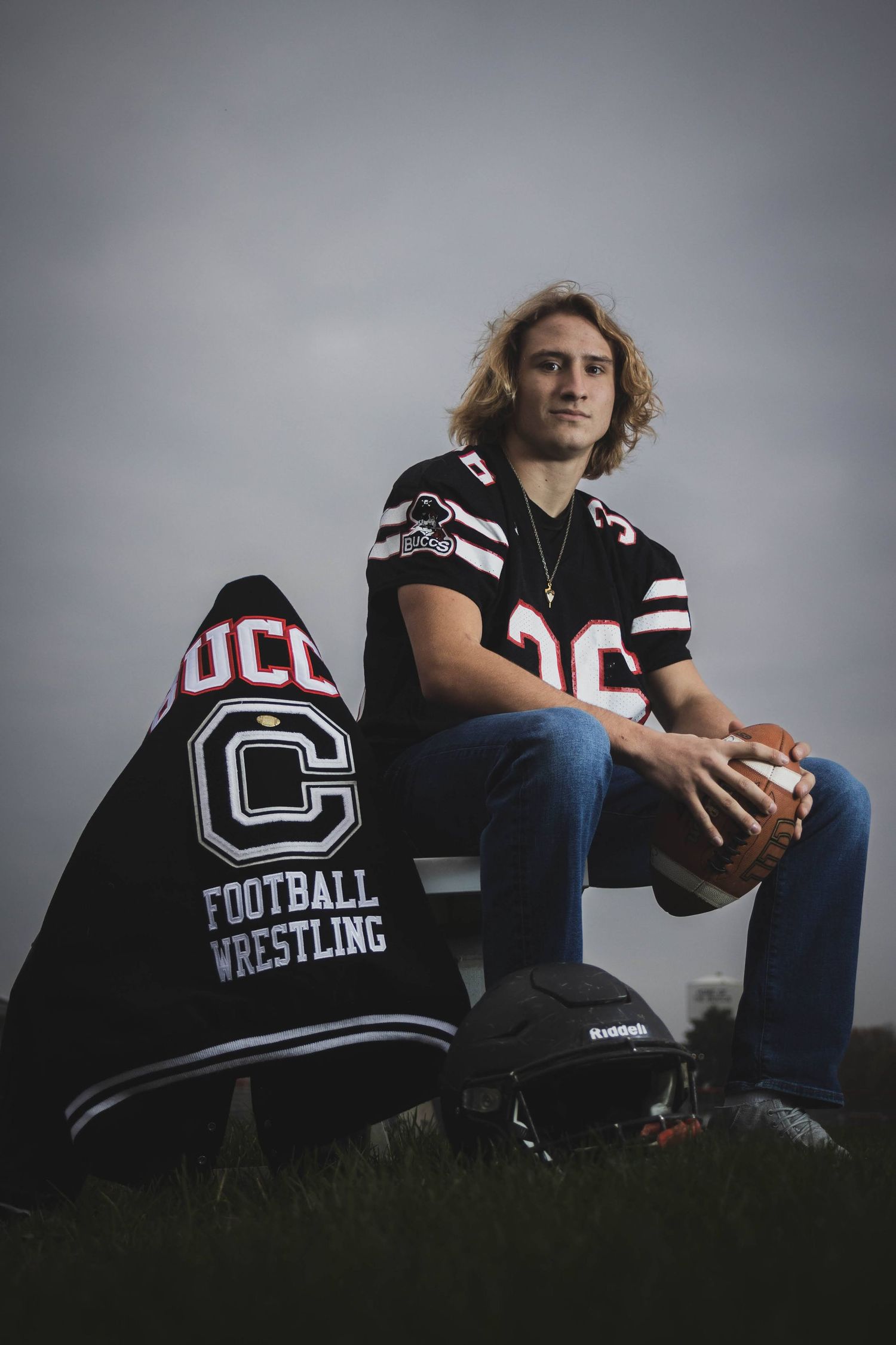 Athletic person in varsity jacket sitting with football against grey background.