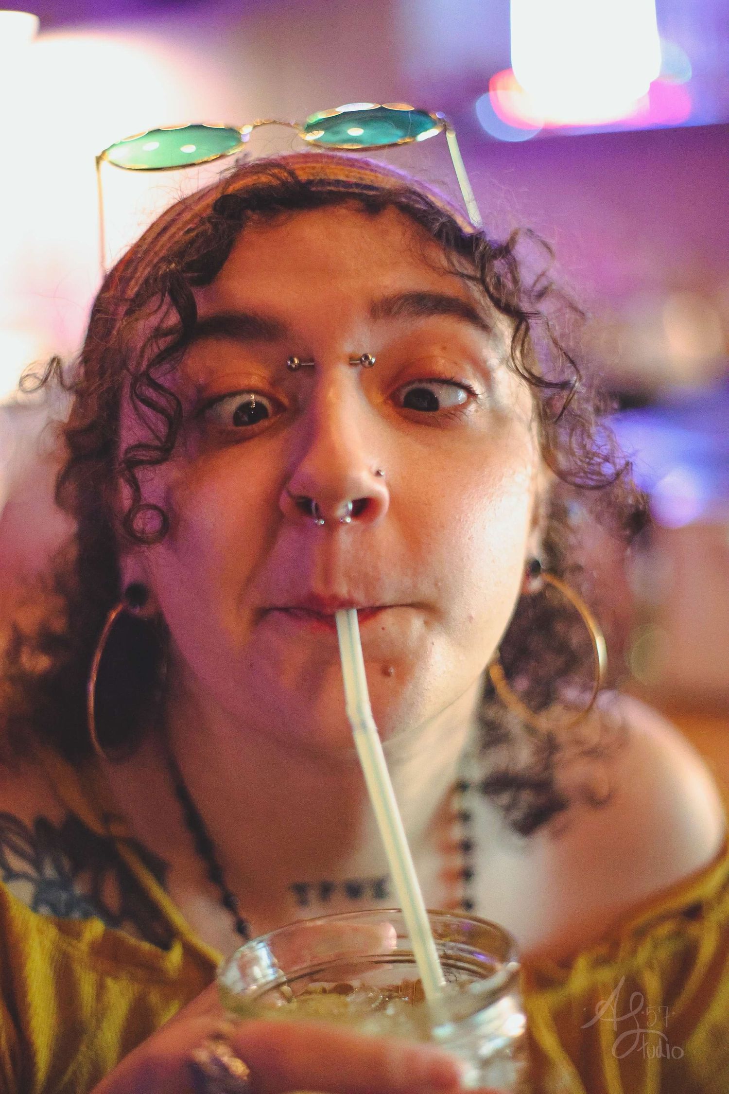 Fun portrait of person with curly hair drinking through straw with sunglasses on head.