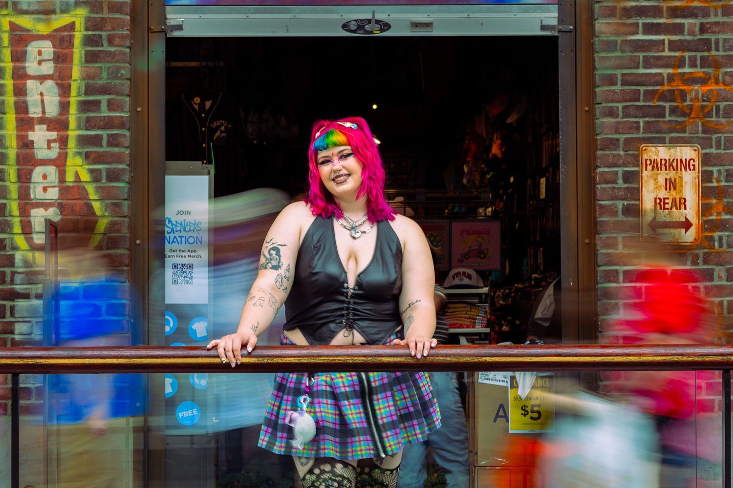 Woman with pink hair in plaid skirt poses at brick building entrance with colorful street scene behind her.