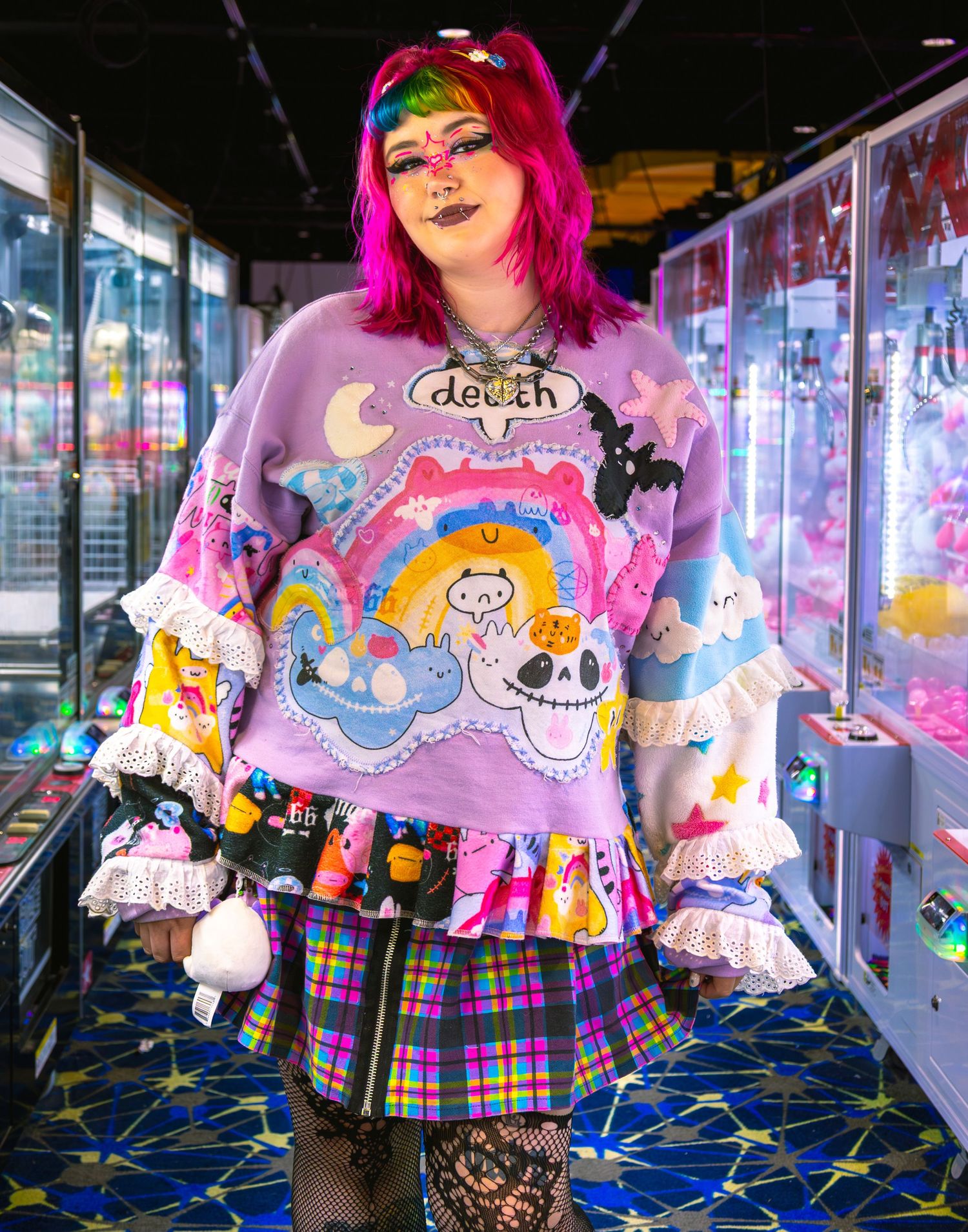 Woman in colorful Harajuku fashion with rainbow hair, graphic sweatshirt, plaid skirt, and fishnet tights in arcade.