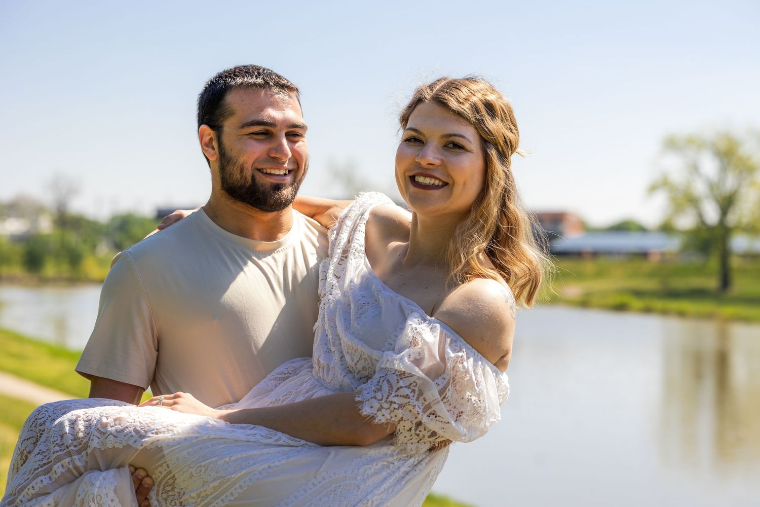 Happy couple posing outdoors near a pond, man carrying woman in white dress on a sunny day.