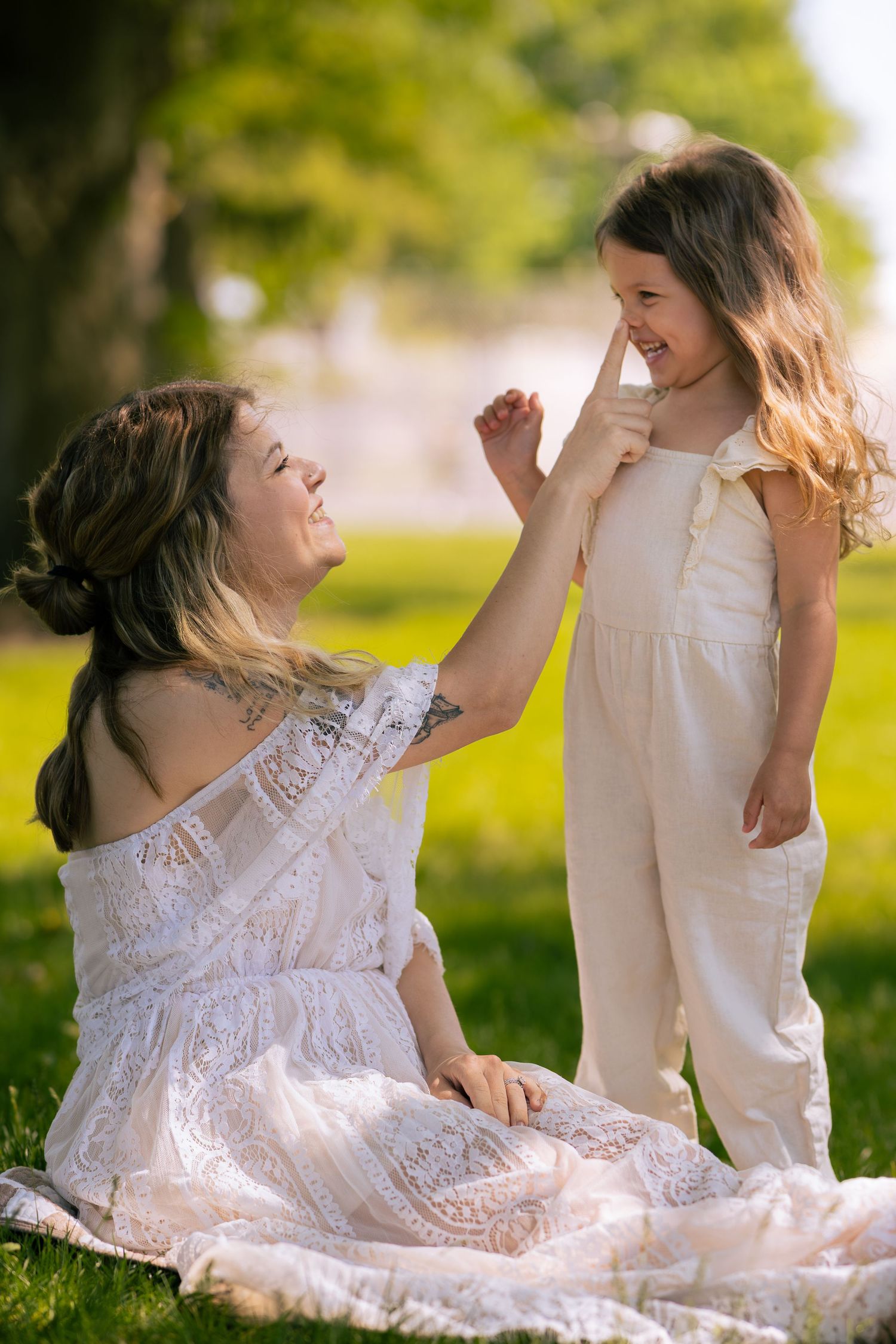 A mother and daughter share a joyful moment in a sunny park, both wearing white outfits, as the girl touches her mom's nose.