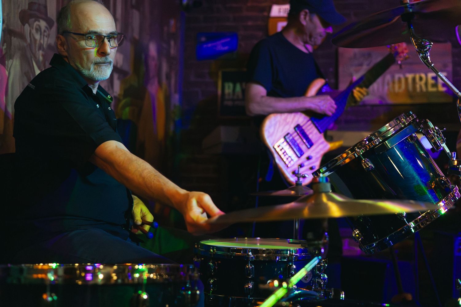 Bald man with glasses playing drums on stage while guitarist performs in background at live music venue.
