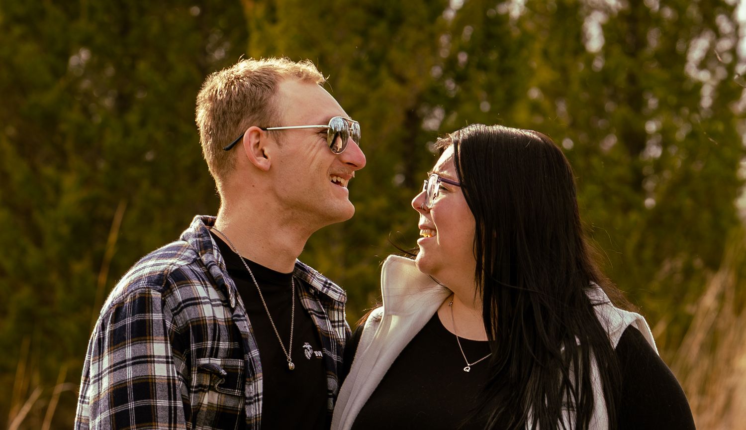 A smiling couple gazes at each other outdoors, man in plaid shirt with sunglasses, woman with long dark hair.