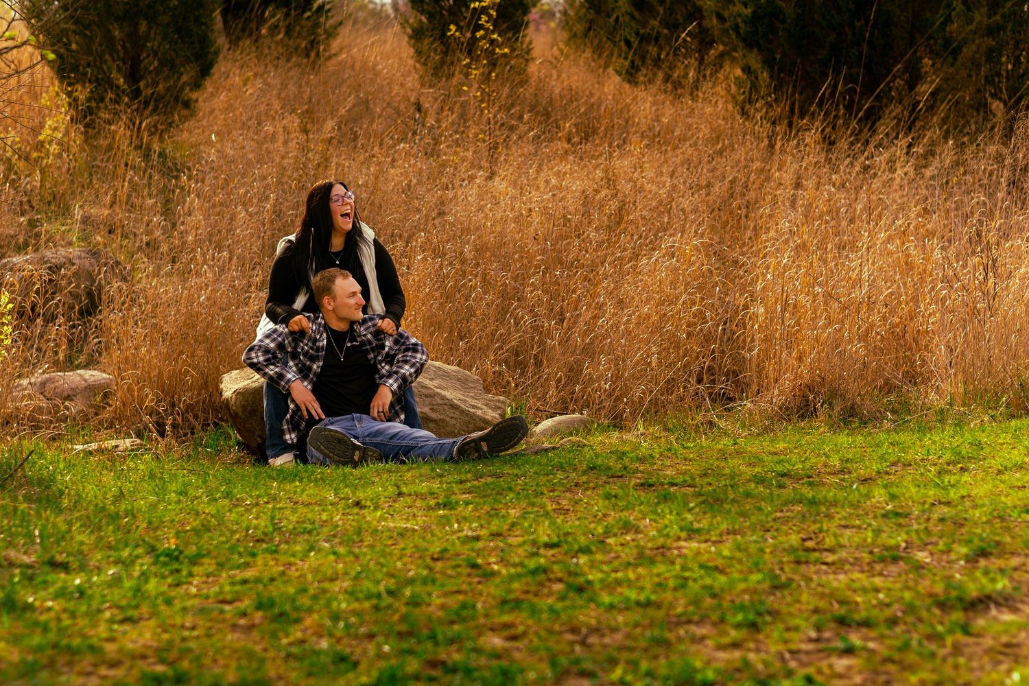 Couple relaxing outdoors in autumn park, woman standing behind seated man near golden tall grass.