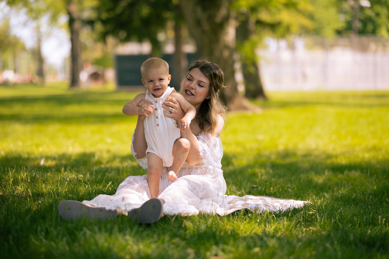 A smiling mother in a white dress sits on grass in a sunny park, joyfully lifting her happy baby in a white onesie.