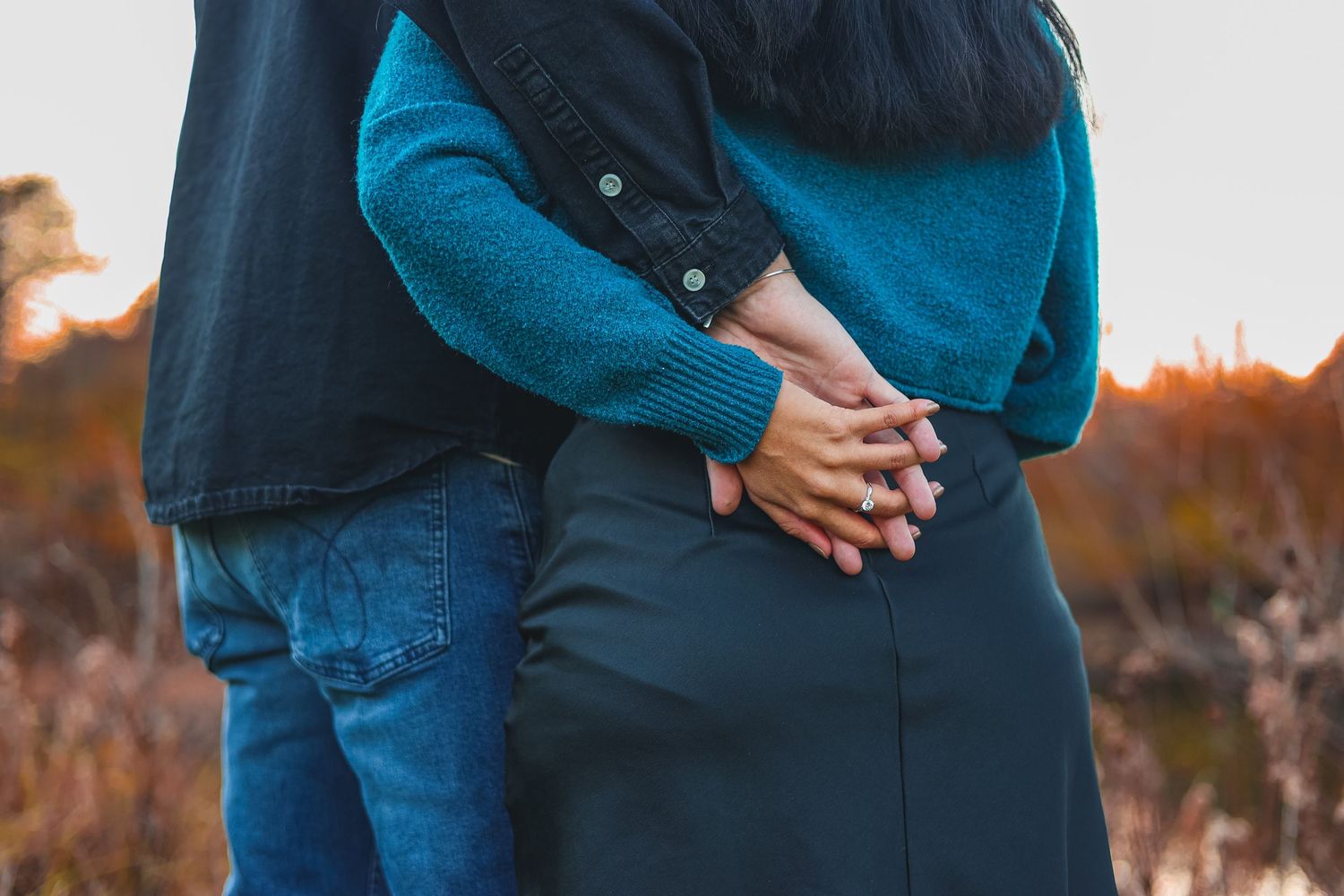 Couple in blue sweaters and jeans embrace during golden hour sunset in a field of tall grass.
