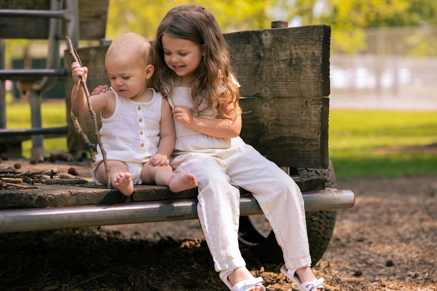 Two young girls sitting together on a playground bench, playing with sticks in a sunny park.